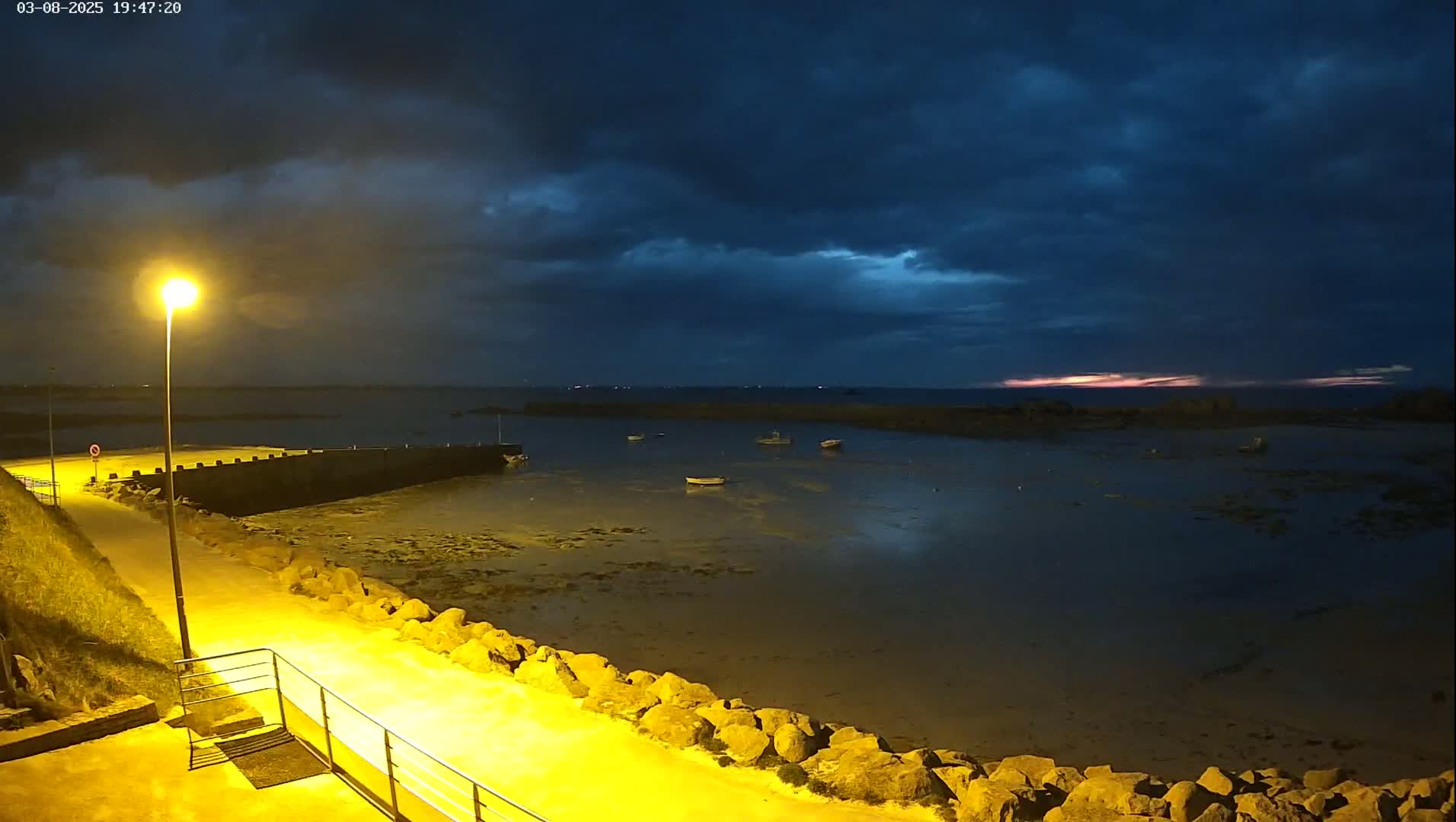 A nighttime view of a calm harbor with several small boats, under a dark, cloudy sky showing a faint hint of pink on the horizon.