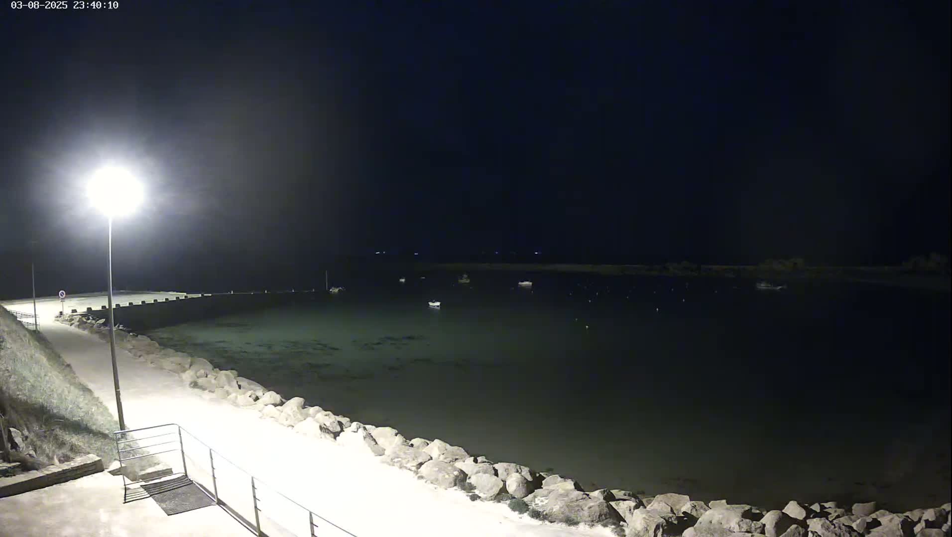 A nighttime view of a calm body of water with several boats moored in a small harbor, a lit pathway leading to the water's edge, and a rocky shoreline.