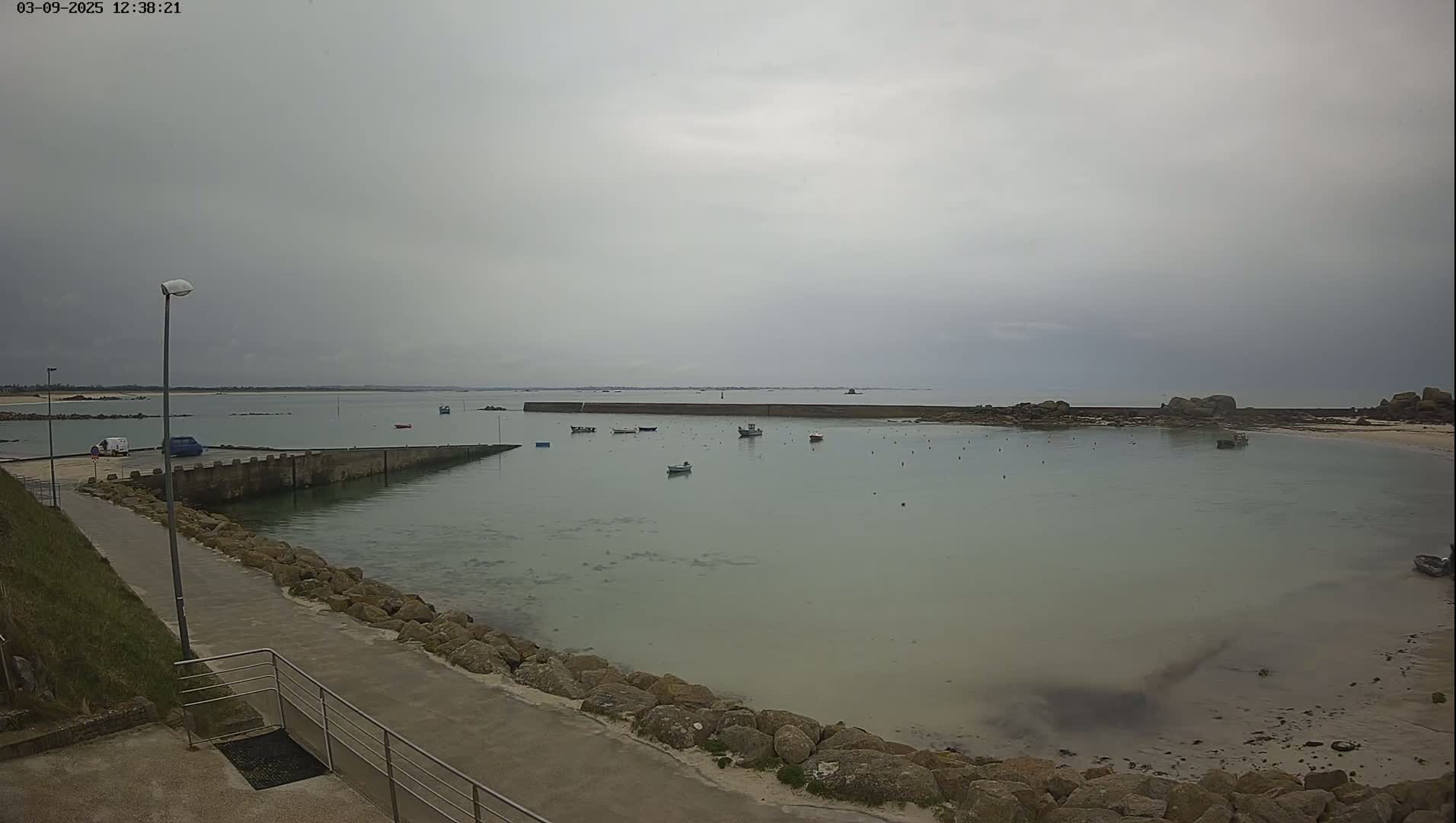 A calm, overcast day reveals a small harbor with several boats, a paved walkway, and rocky shores.