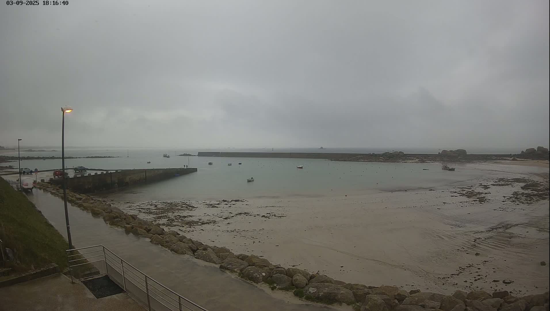 A wet, overcast day reveals a small harbor with several boats at low tide, a stone seawall, and a roadway leading to it.