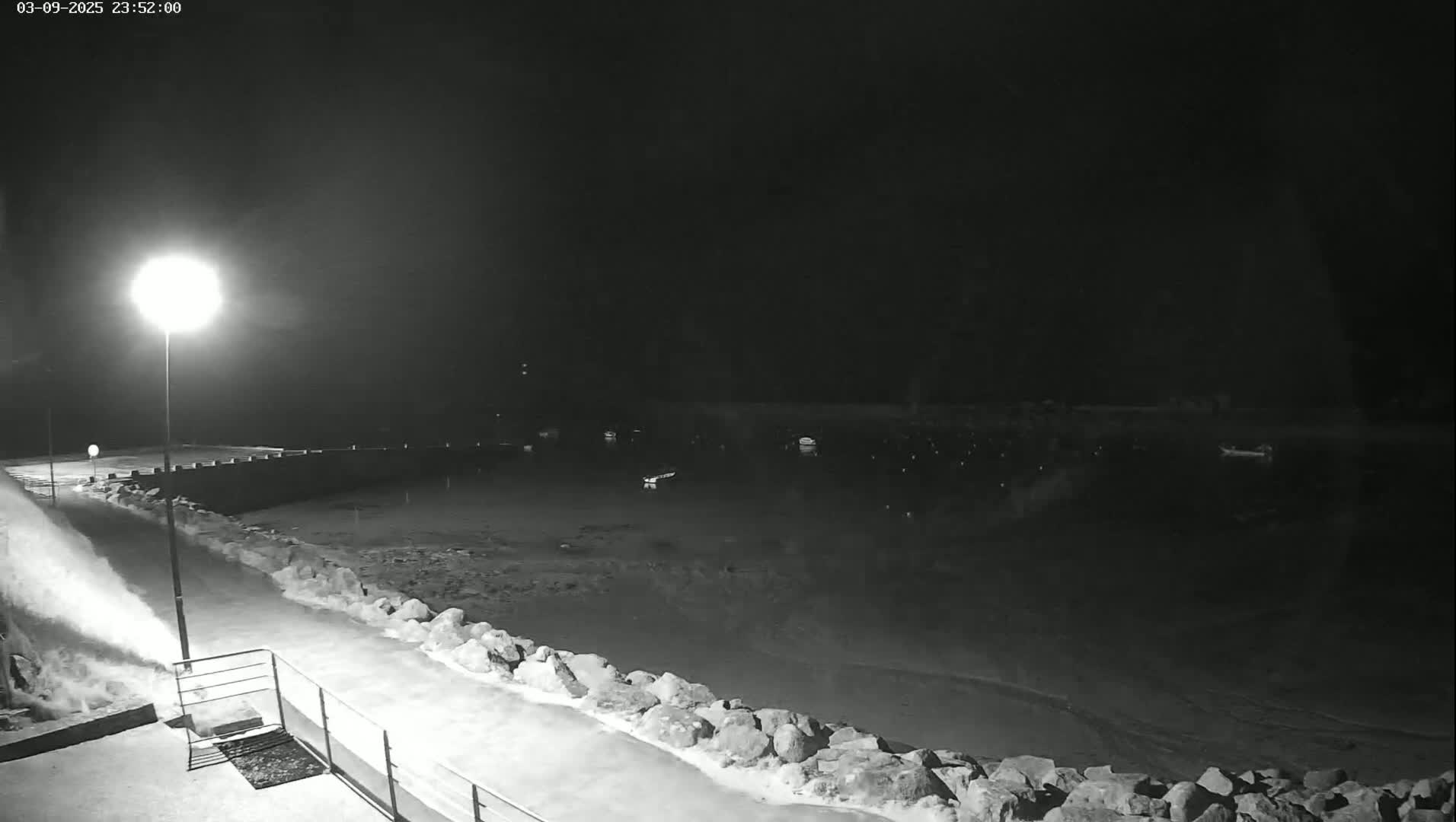 A nighttime, snow-covered harbor scene is illuminated by a single bright lamp, showing a calm body of water with several boats and a snow-removing machine spraying snow.