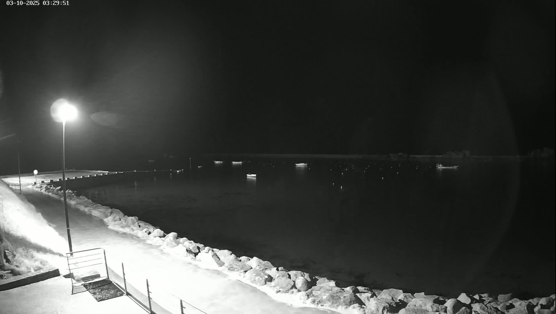 A nighttime view shows a snow-covered walkway next to a calm body of water where several small boats are moored.