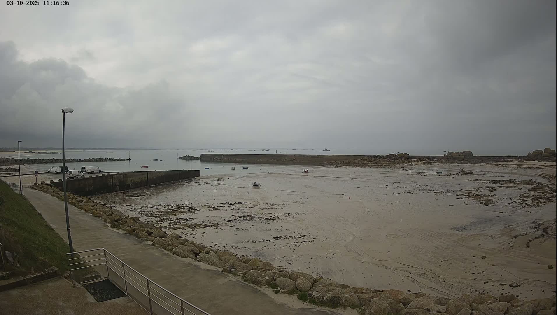 A mostly cloudy day reveals a low-tide coastal scene with a small harbor, a few boats, and a paved walkway leading to a rocky shore.