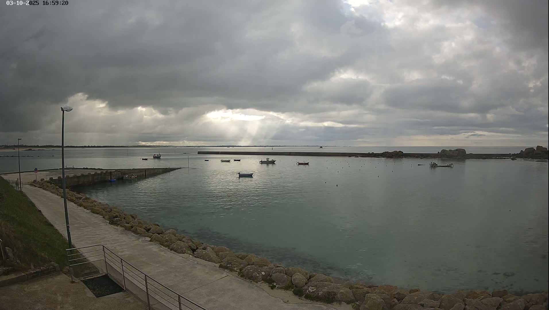 A calm harbor with several small boats under a cloudy sky, with sunbeams breaking through the clouds.