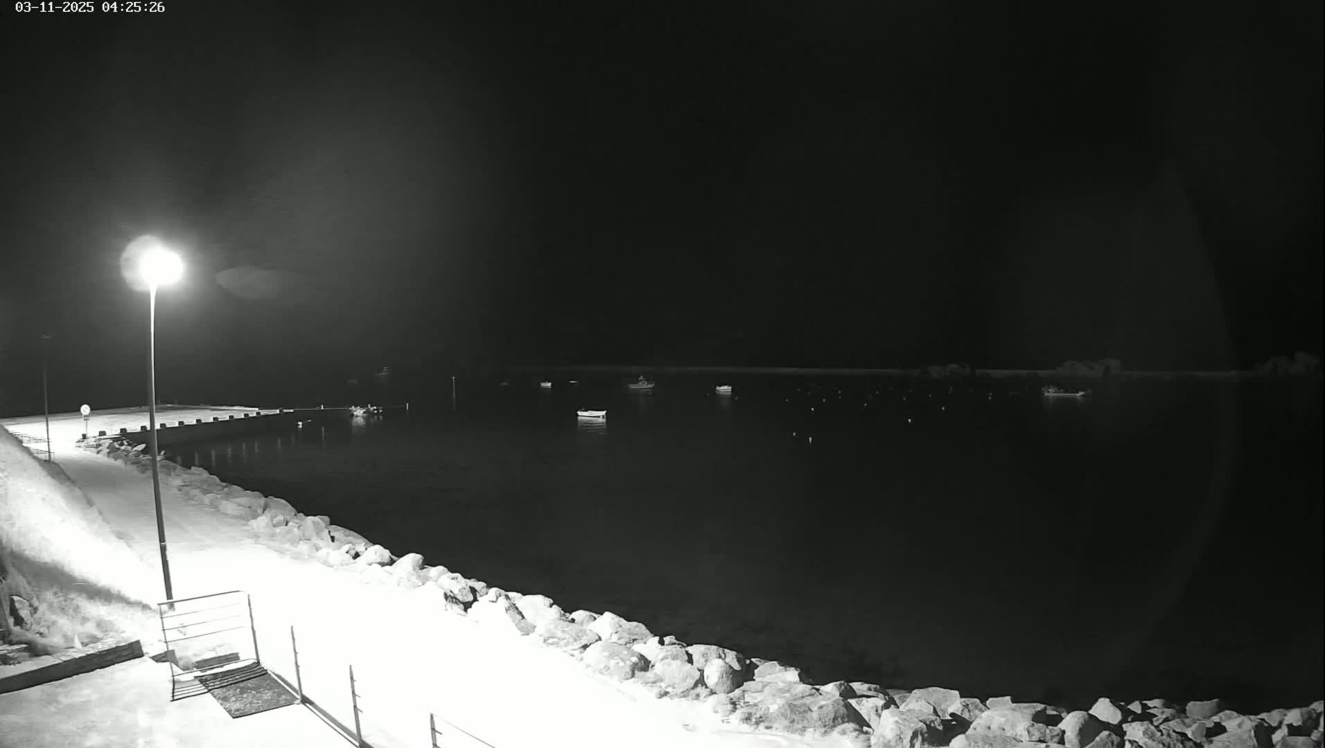 A nighttime, snow-covered waterfront scene shows several small boats moored in a calm harbor, illuminated by a single lamppost on a pier.