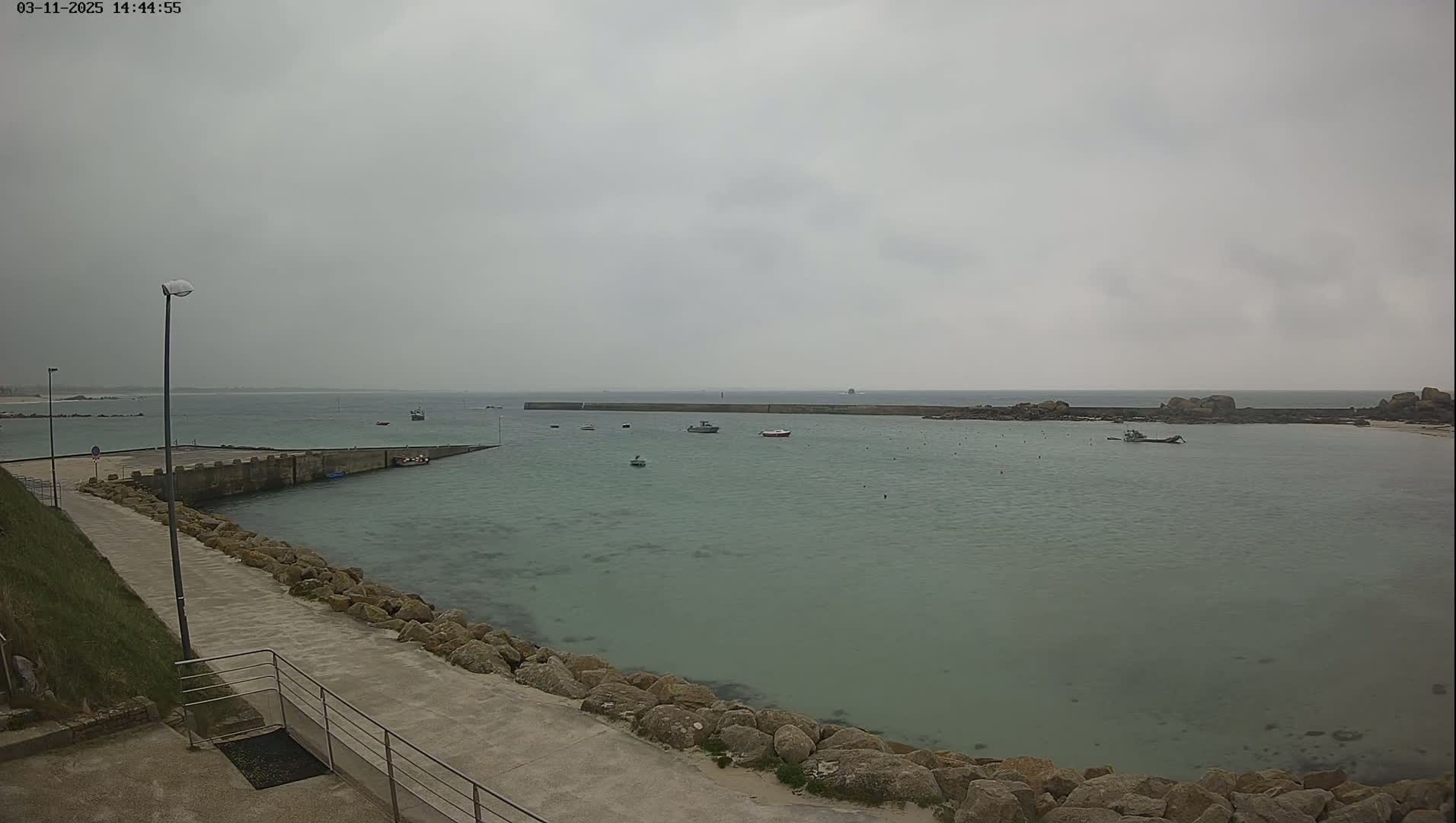 A calm, overcast day shows a harbor with several small boats, a stone walkway, and rocky shores.