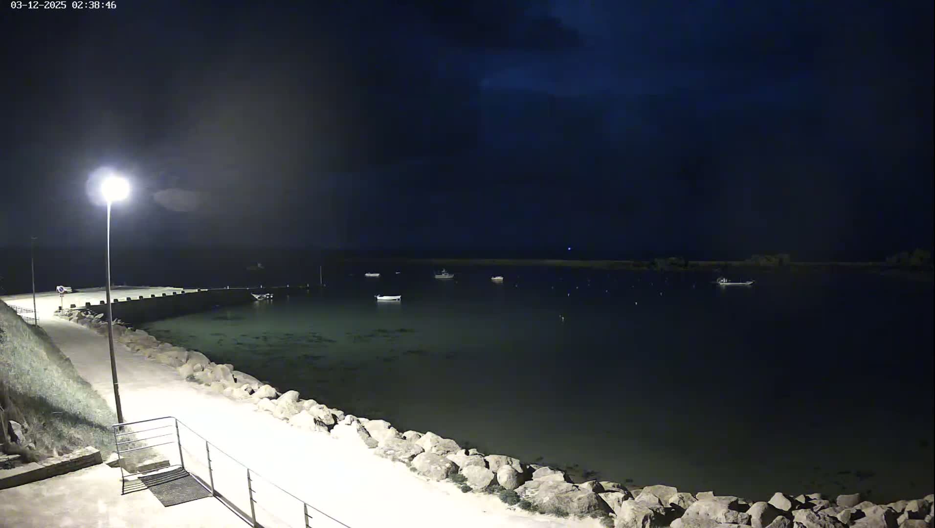 A nighttime view of a calm body of water with several small boats moored, a lit walkway along a rocky shoreline, and a single lamppost.