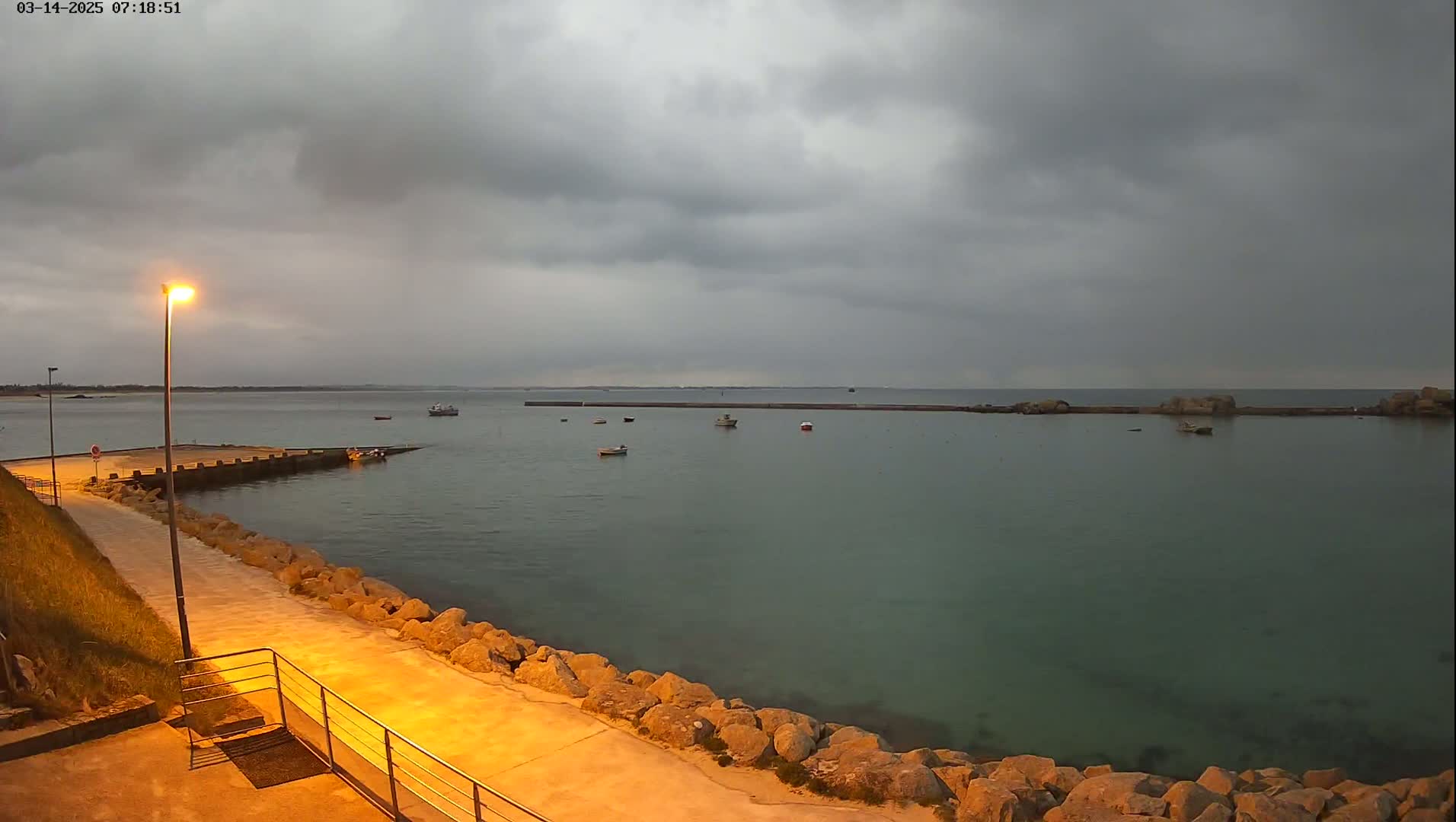 A calm harbor with several small boats under a cloudy, overcast sky, lit by a single lamppost on a paved walkway.