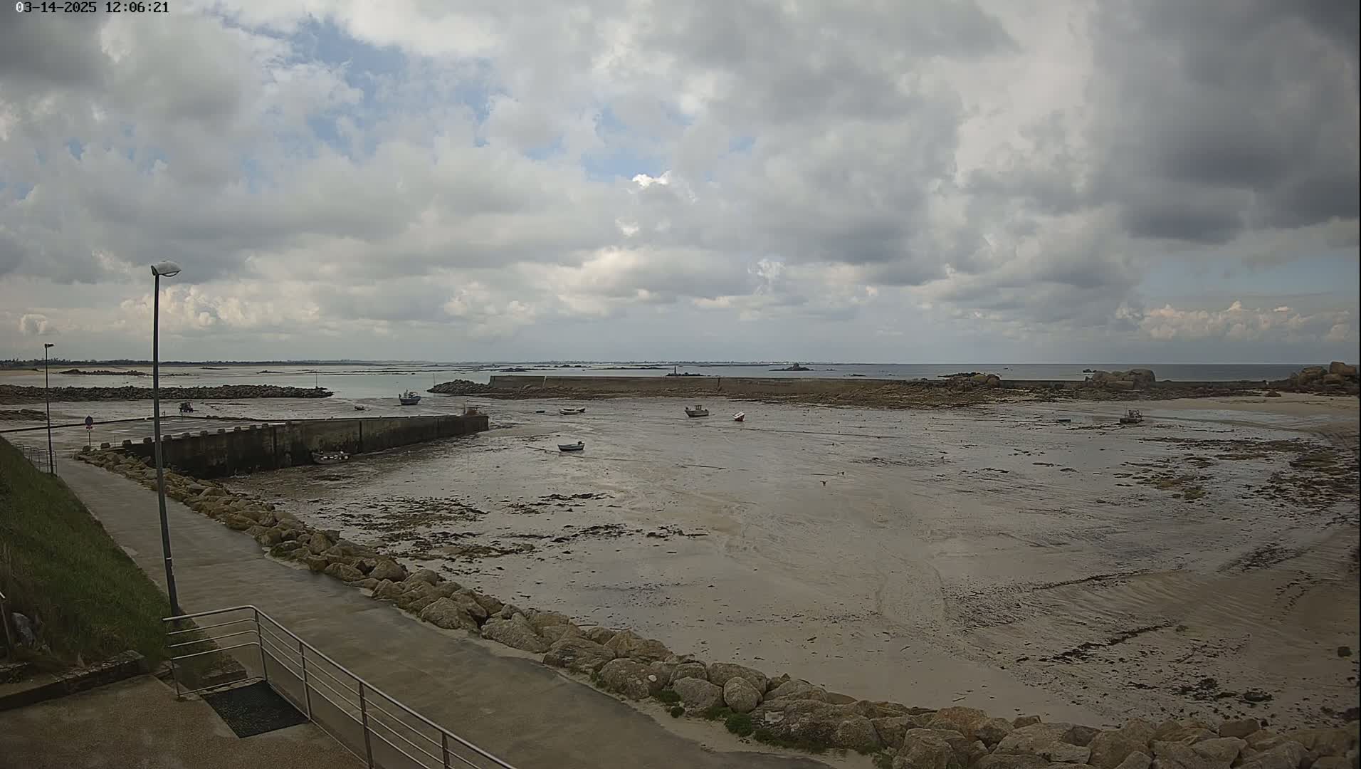A mostly cloudy day reveals a small harbor with several small boats at low tide, a stone seawall, and a paved walkway leading to it.