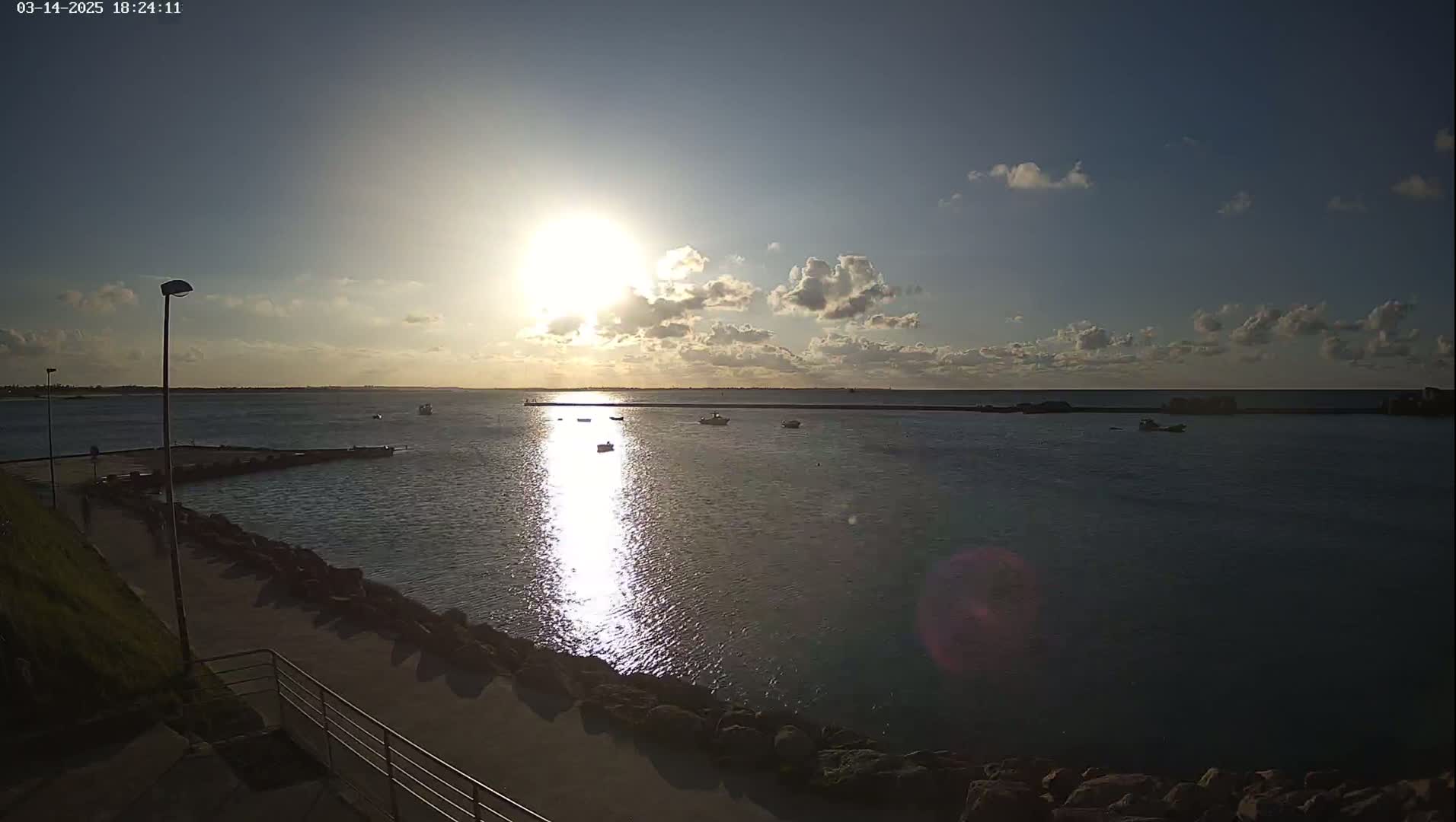 A partly cloudy sunset over calm water with several small boats, viewed from a paved walkway next to a rocky shoreline.