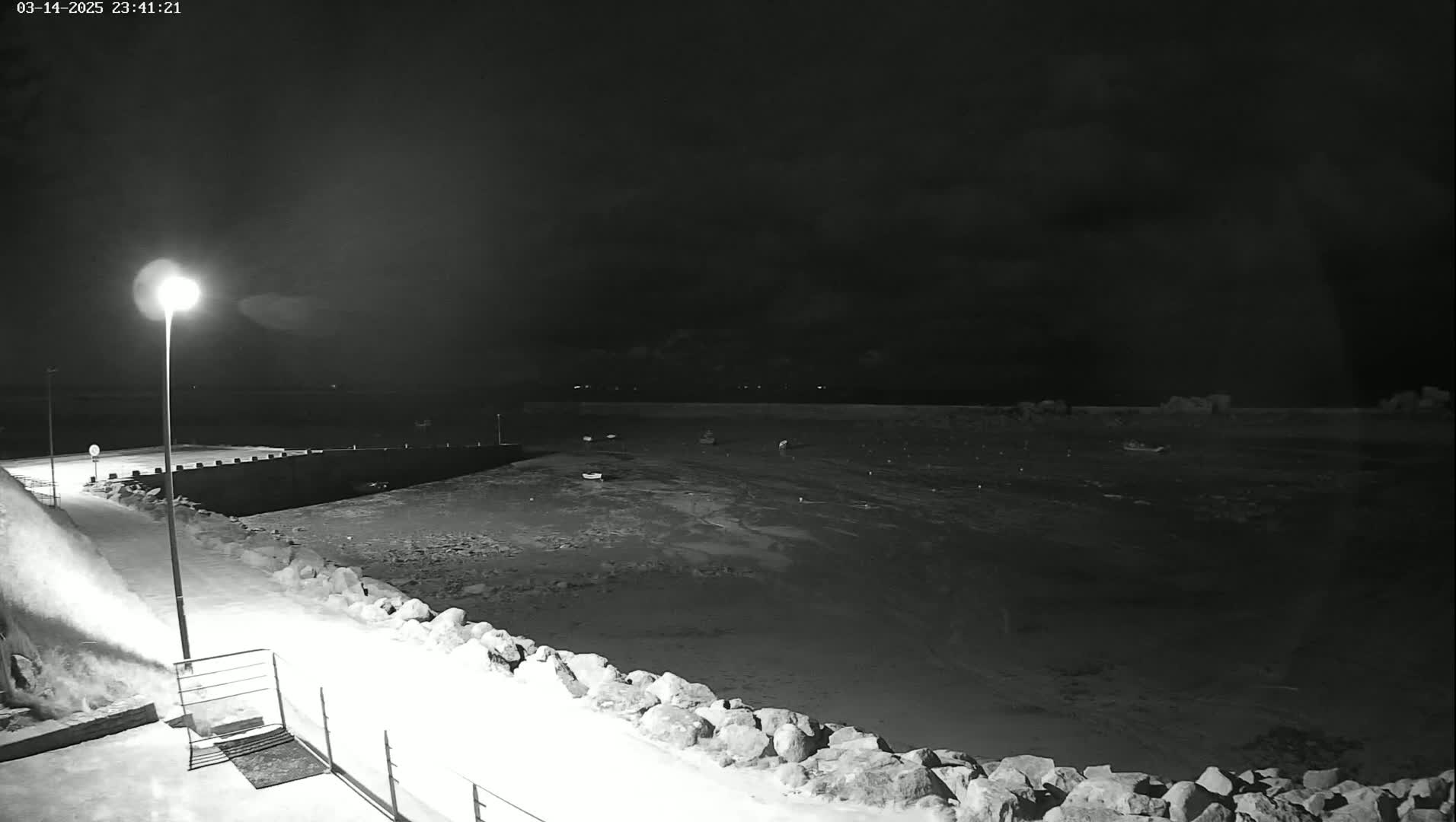 A nighttime, snowy view of a harbor with several small boats at rest under a cloudy sky.