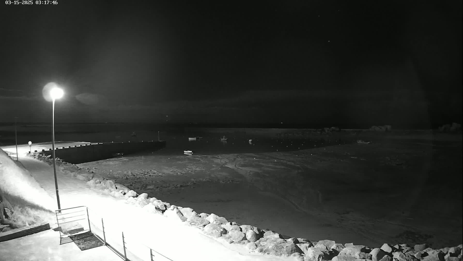A nighttime, snow-covered harbor scene shows several small boats moored in calm water, illuminated by a single lamppost on a rocky shoreline.
