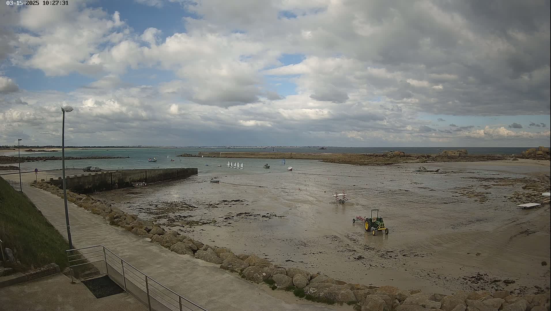 A partly cloudy day reveals a small harbor with several sailboats, a tractor, and exposed tidal flats, all situated near a paved walkway and a rocky shoreline.