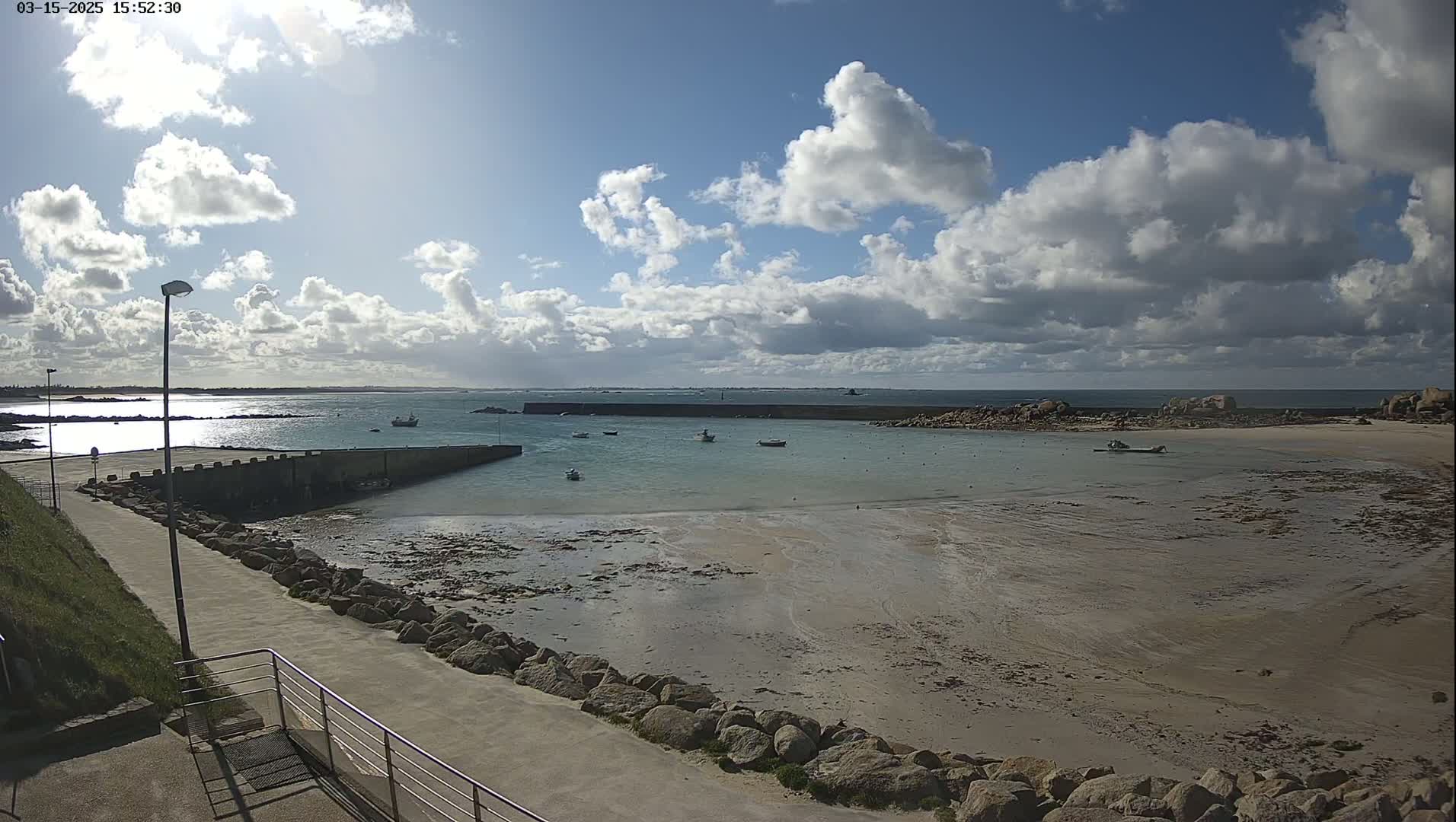 A partly sunny day reveals a rocky, sandy cove with several small boats moored in calm water, a stone jetty, and a paved walkway leading to it.