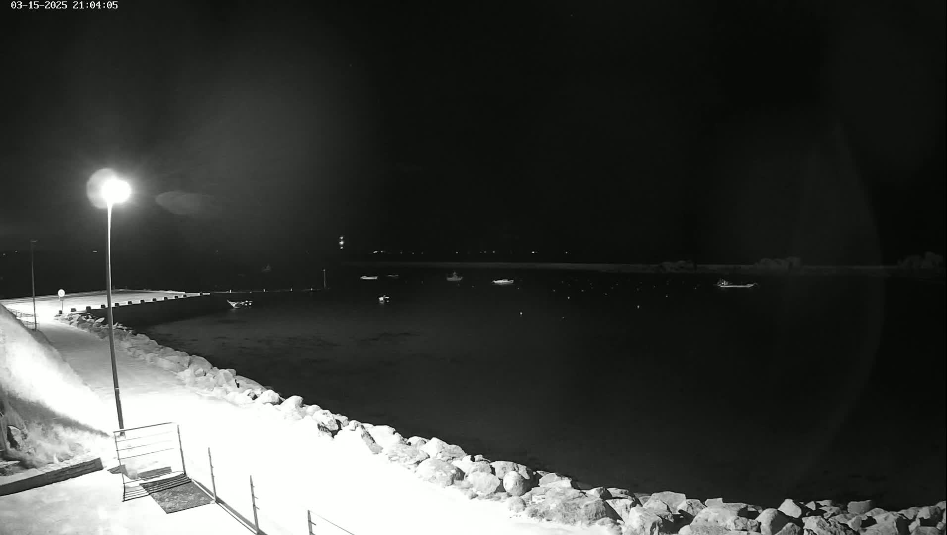 A snow-covered rocky shoreline at night is illuminated by a single lamppost, with several boats visible in the dark water beyond.