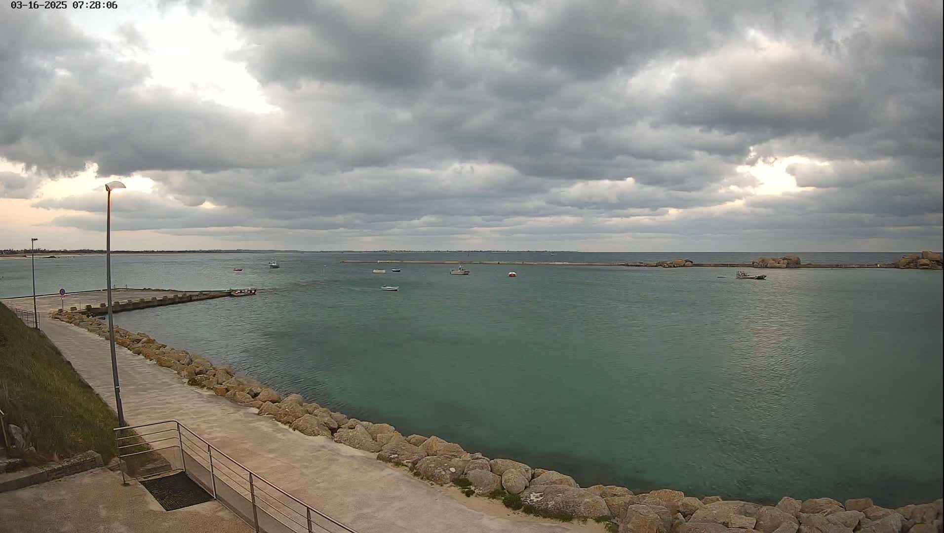 A calm, teal-colored body of water is partially enclosed by a rocky shore with a paved walkway and a few small boats scattered across its surface under a cloudy sky.