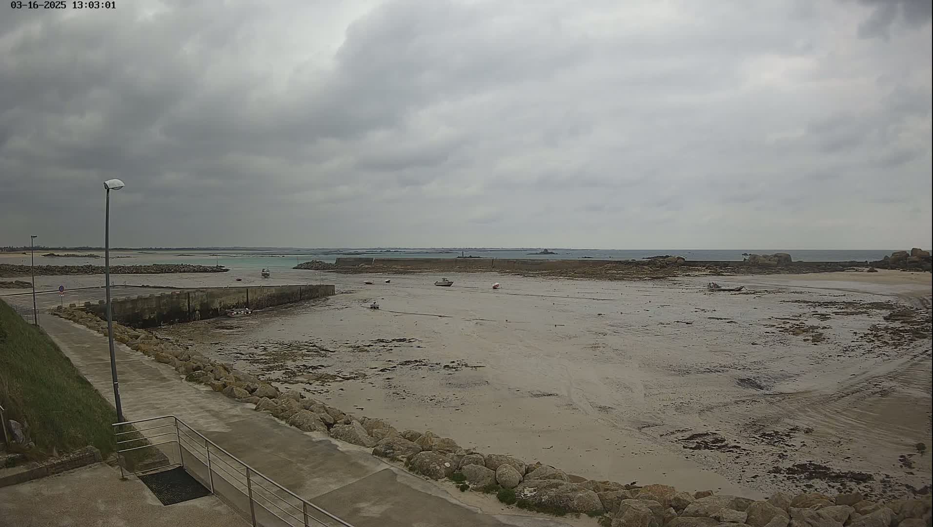 A mostly overcast sky hangs over a low-tide, sandy harbor with a few small boats and a paved walkway leading to it.