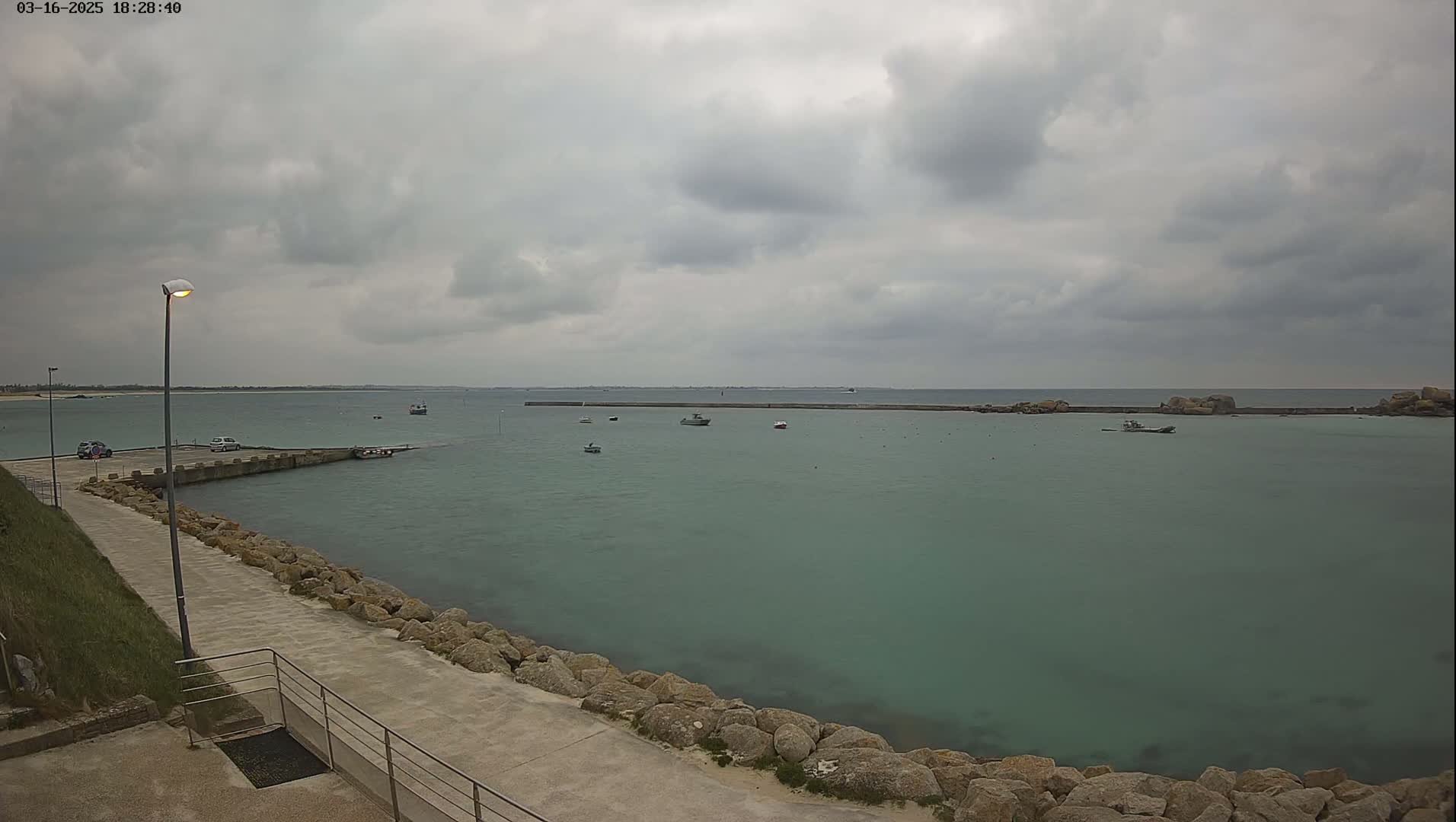 A calm, overcast day shows a harbor with several small boats, a stone seawall, and a paved walkway leading to a parking area.