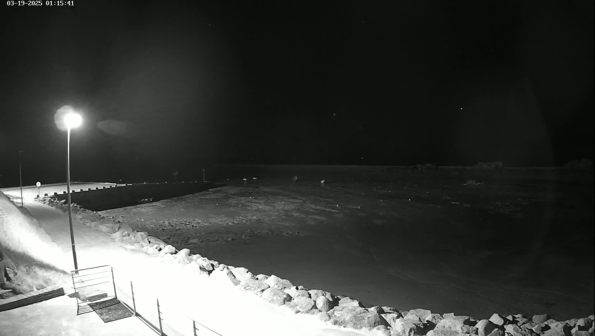 A snow-covered waterfront at night, illuminated by a single lamppost, shows a dark body of water partially frozen over.