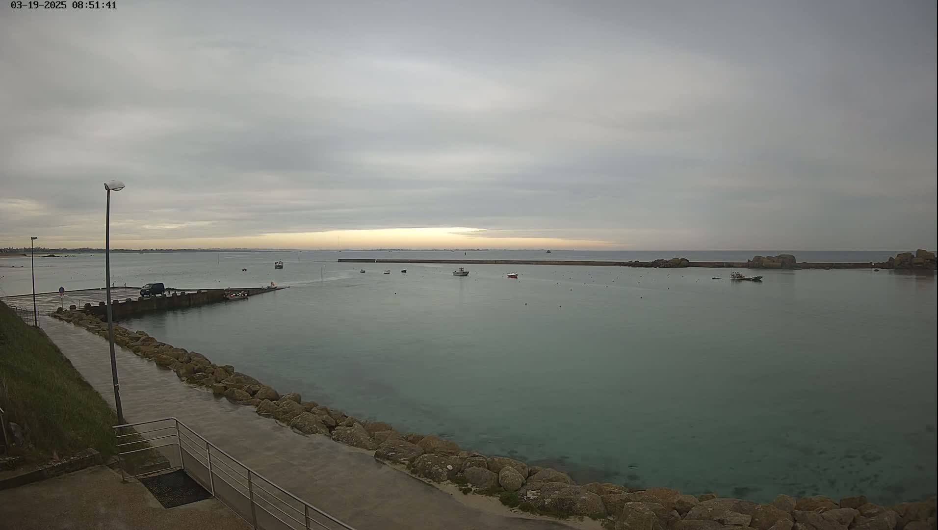 A calm, overcast day at a harbor shows several small boats moored in the water near a rocky shoreline, with a paved walkway leading to a small parking area.