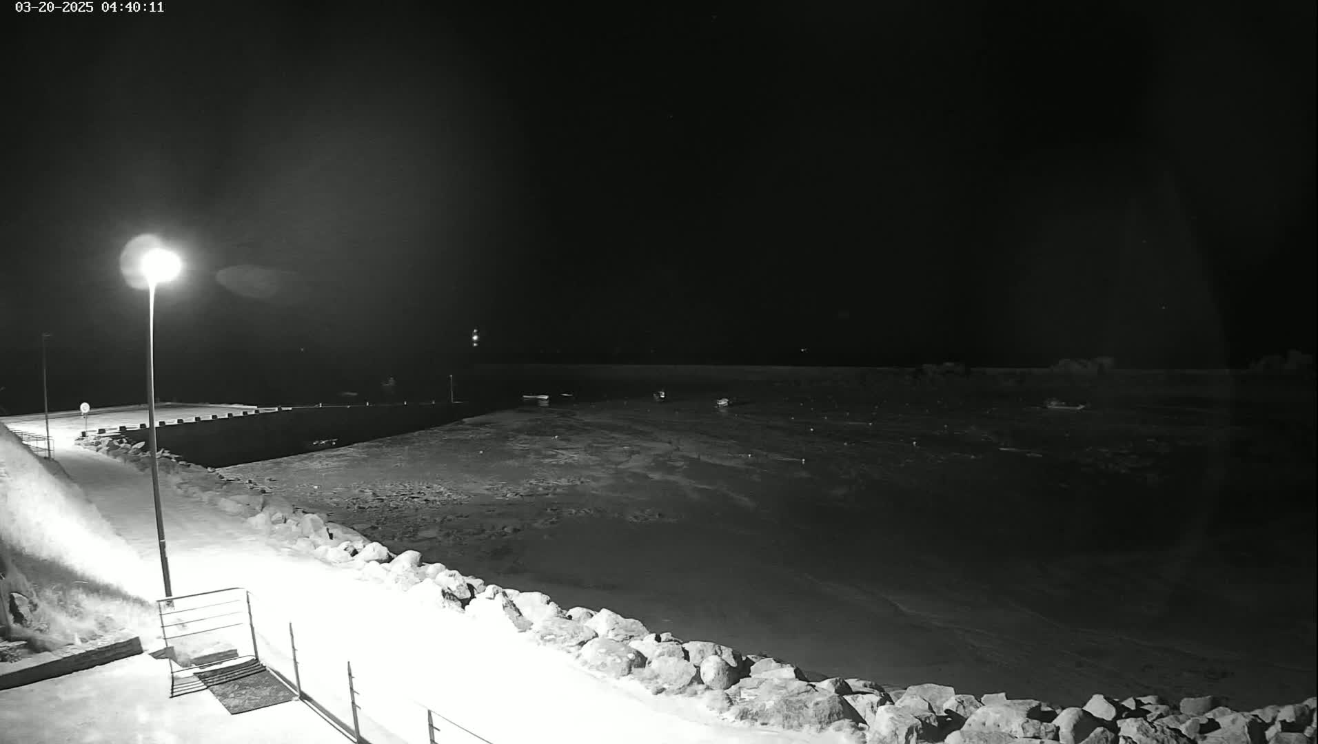 A snow-covered harbor at night, illuminated by a single lamppost, shows several boats moored in the dark water.