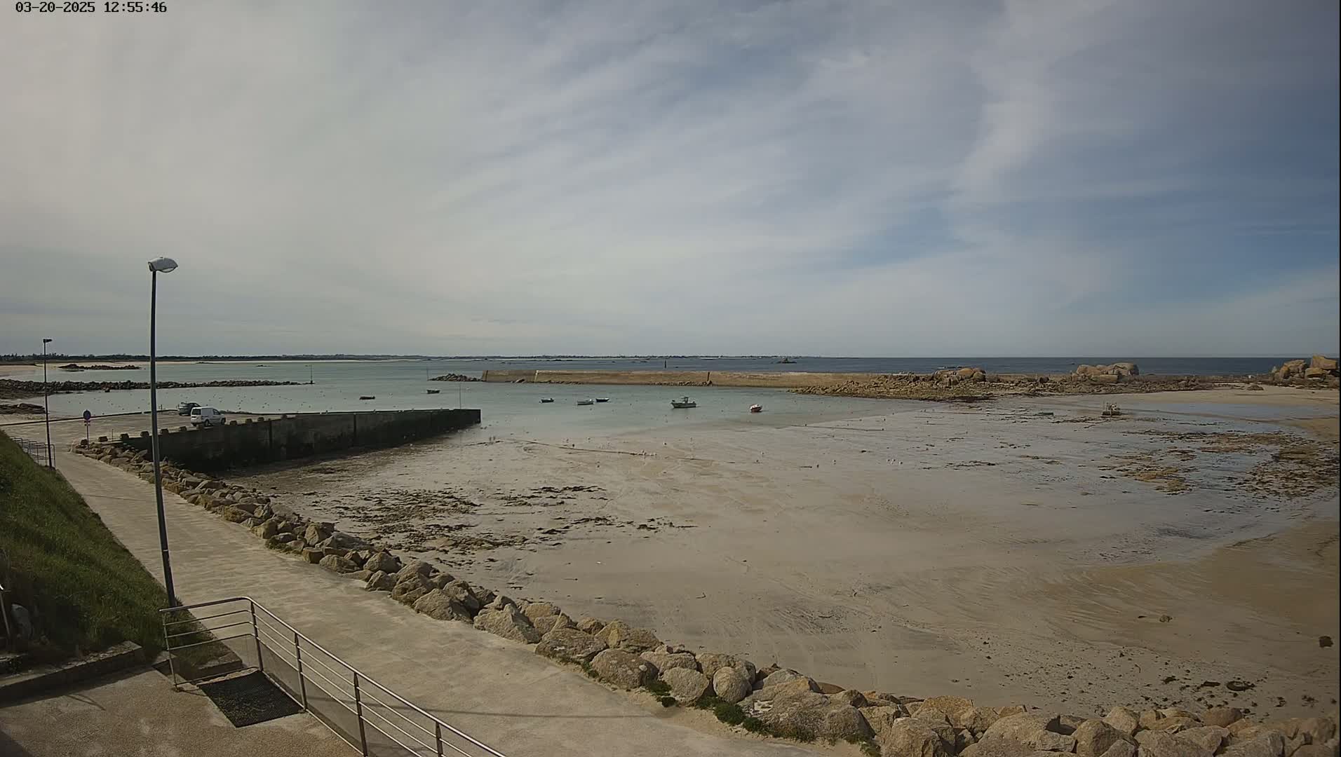 A mostly sunny day reveals a small harbor with several boats, a sandy beach exposed at low tide, and a paved walkway lined with rocks leading to the water.