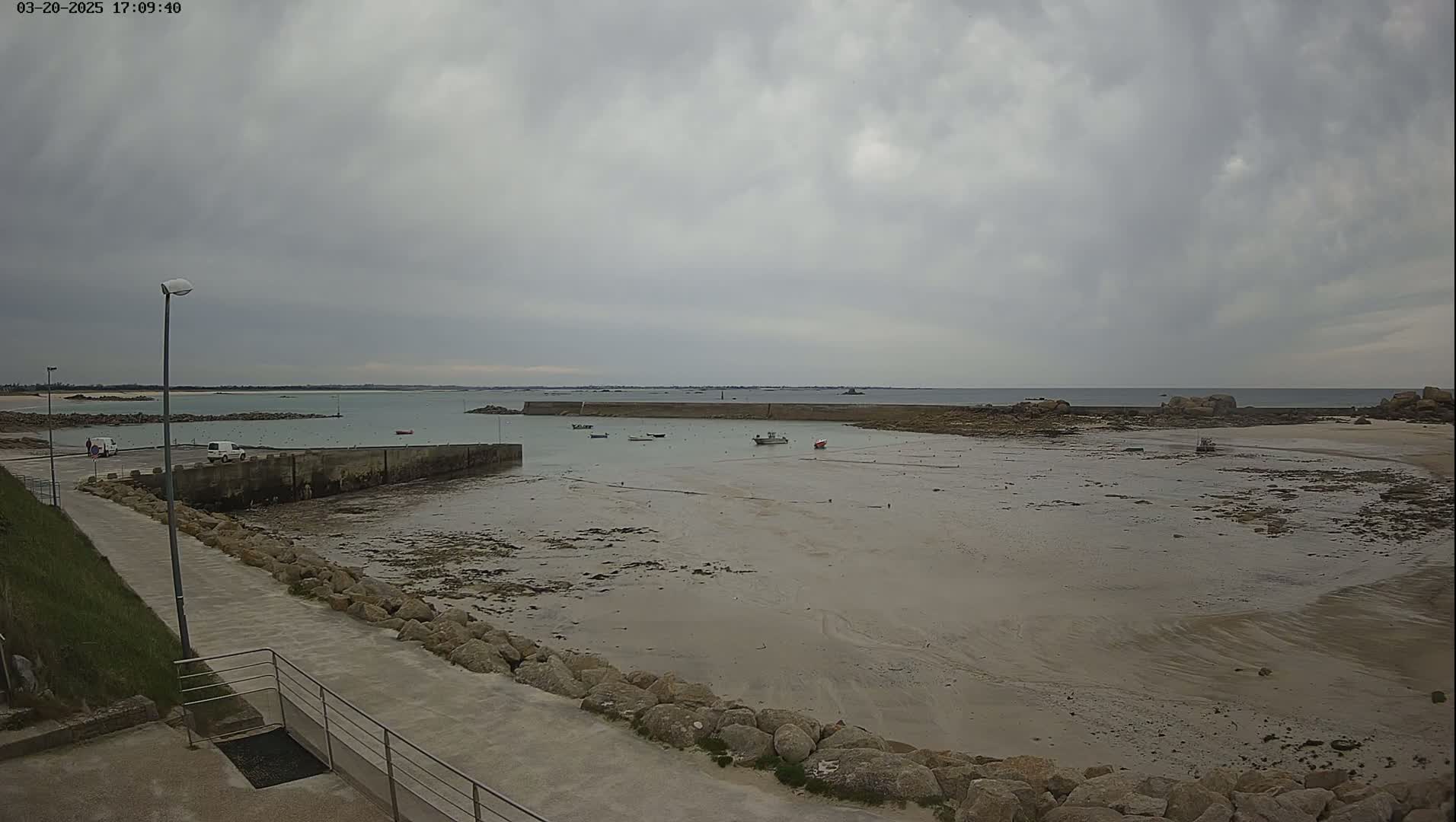 A low-tide scene shows a small harbor with several boats, a paved walkway, and a rocky shoreline under an overcast sky.