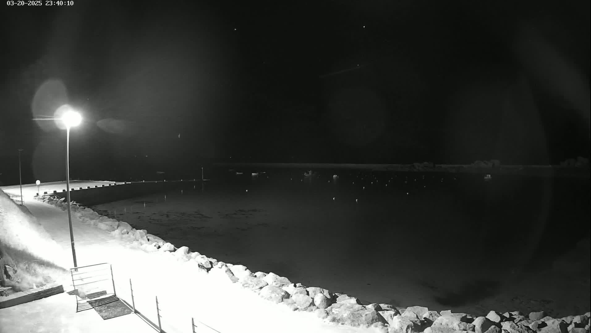 A snow-covered harbor at night is illuminated by a single lamppost, with several boats visible in the calm water.