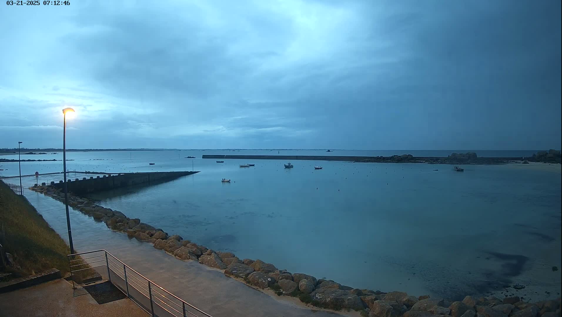 A calm, overcast harbor at dawn shows several small boats moored in shallow, clear water, with a stone walkway leading to a pier.