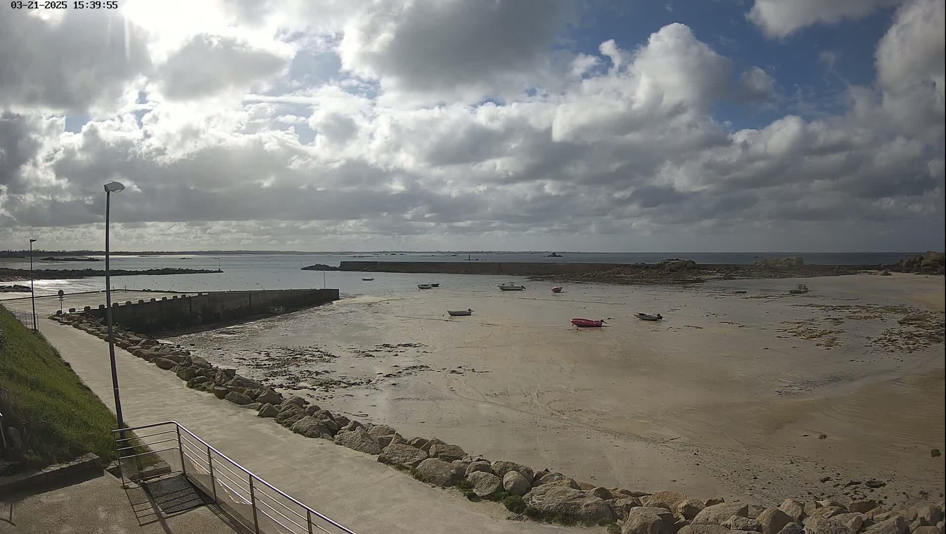 A mostly sunny day with partly cloudy skies shows a sandy beach with several small boats at low tide near a stone seawall and paved walkway.