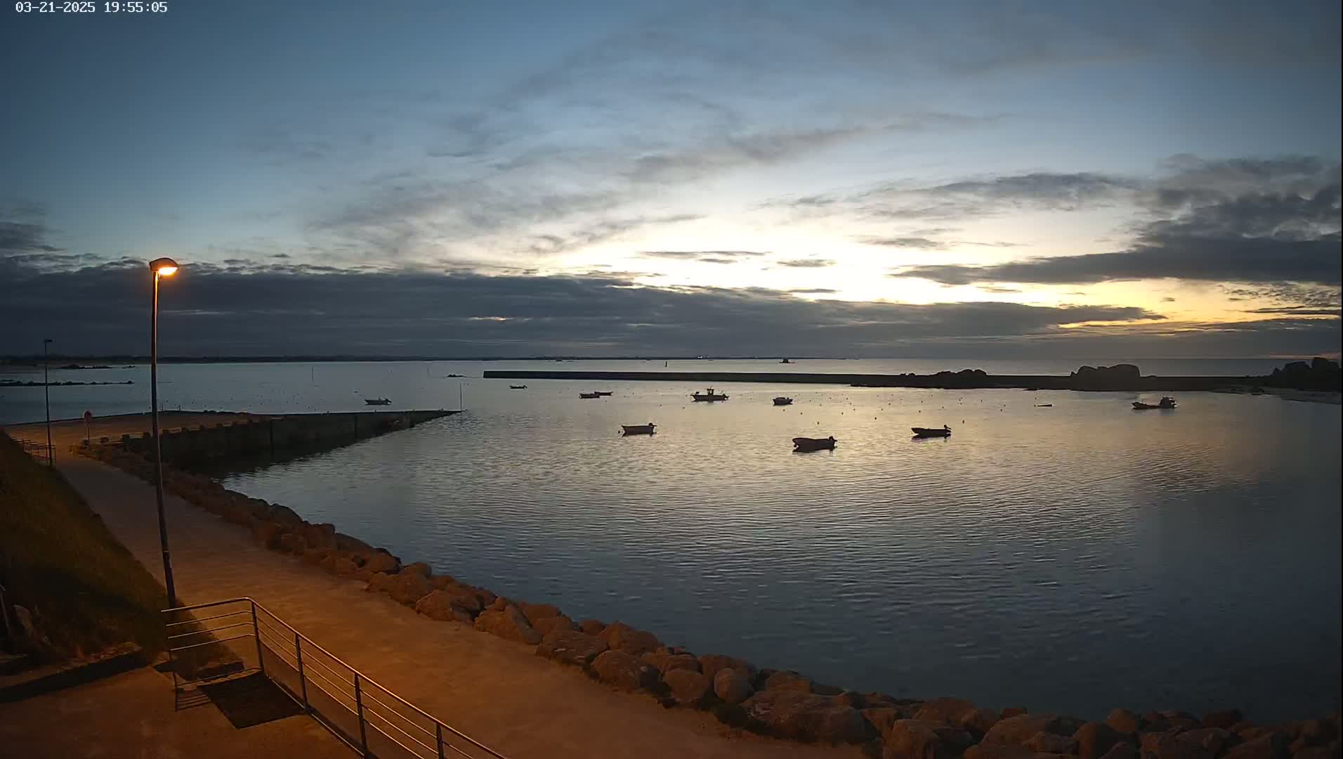 A calm harbor at dusk, with several small boats moored, is seen from a paved walkway next to a rocky shoreline under a partly cloudy sky.