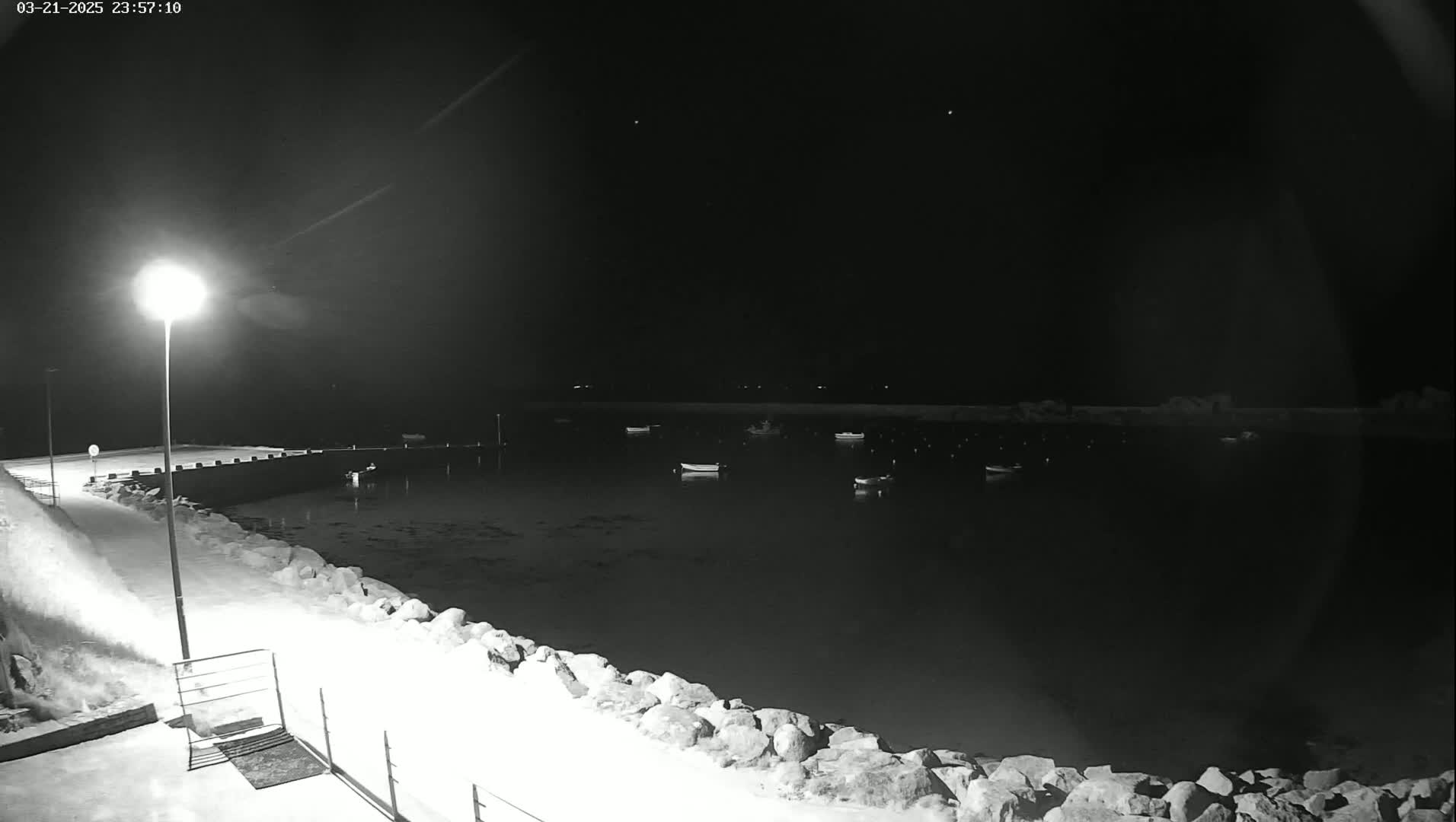 A nighttime, snow-covered harbor scene shows several small boats moored in calm water under a dark sky illuminated by a single lamppost.