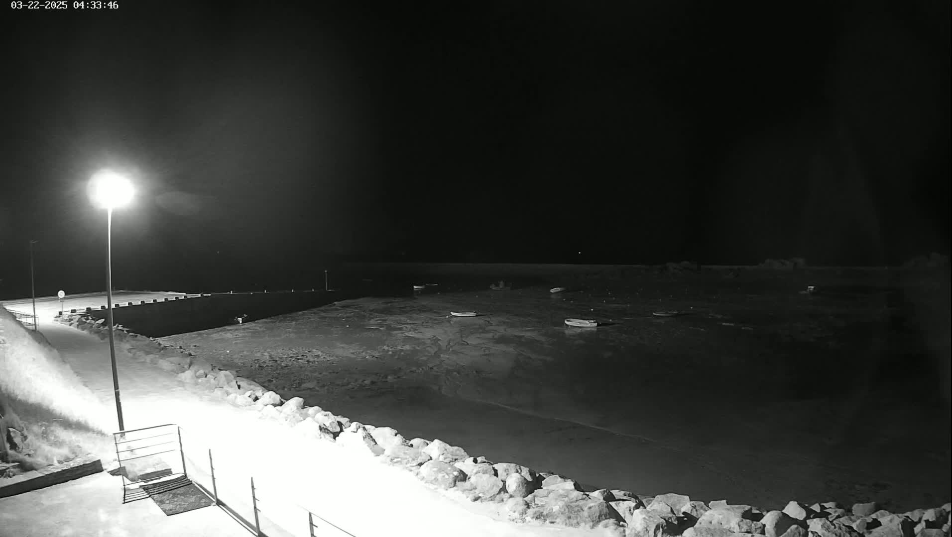 A nighttime, snow-covered shoreline with a few small boats in calm water is illuminated by a single lamppost.