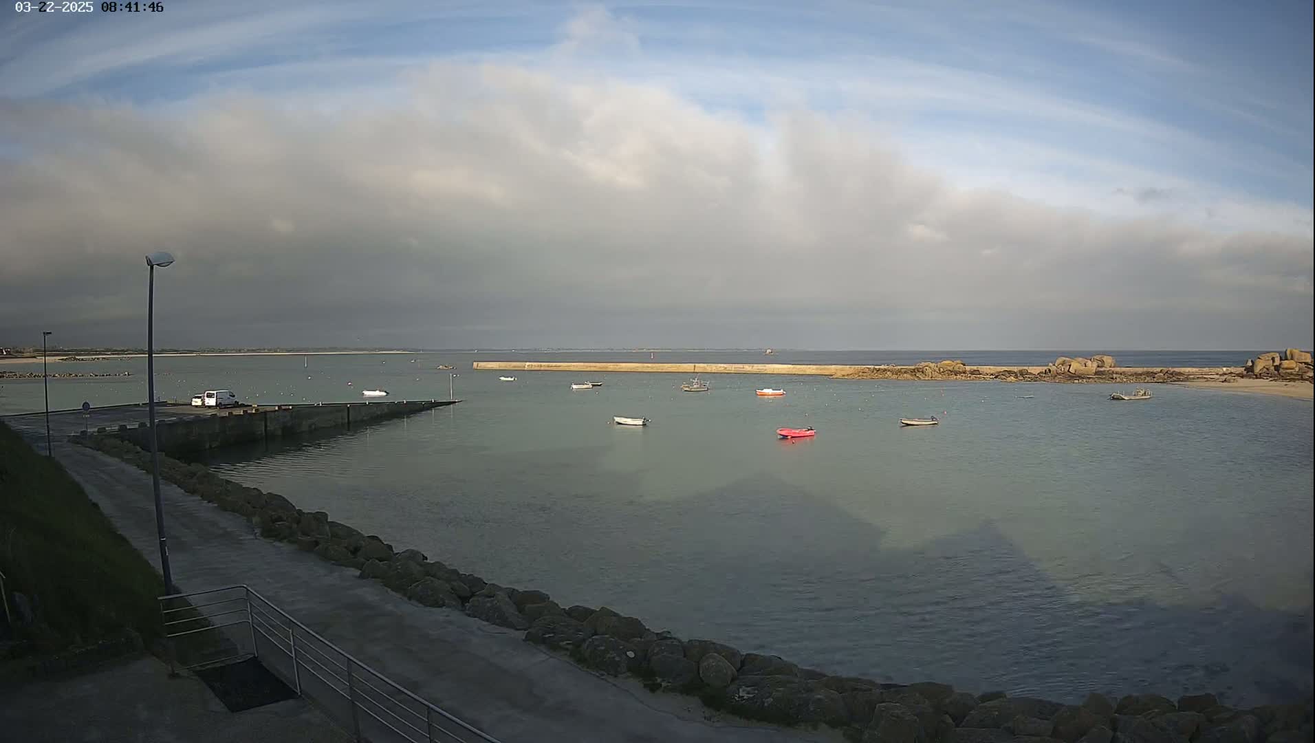 A calm harbor with several small boats is viewed from a paved area on an overcast day.