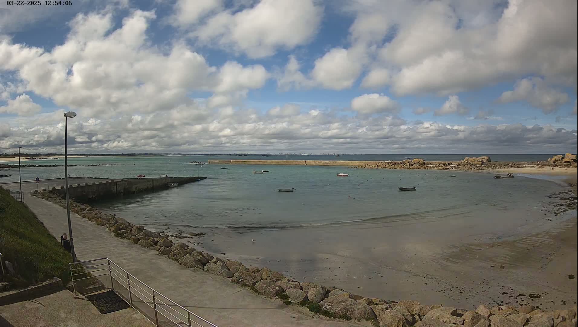 A calm, partly cloudy day reveals a small harbor with several boats at rest, a sandy beach, and a rocky shoreline extending to a calm sea.