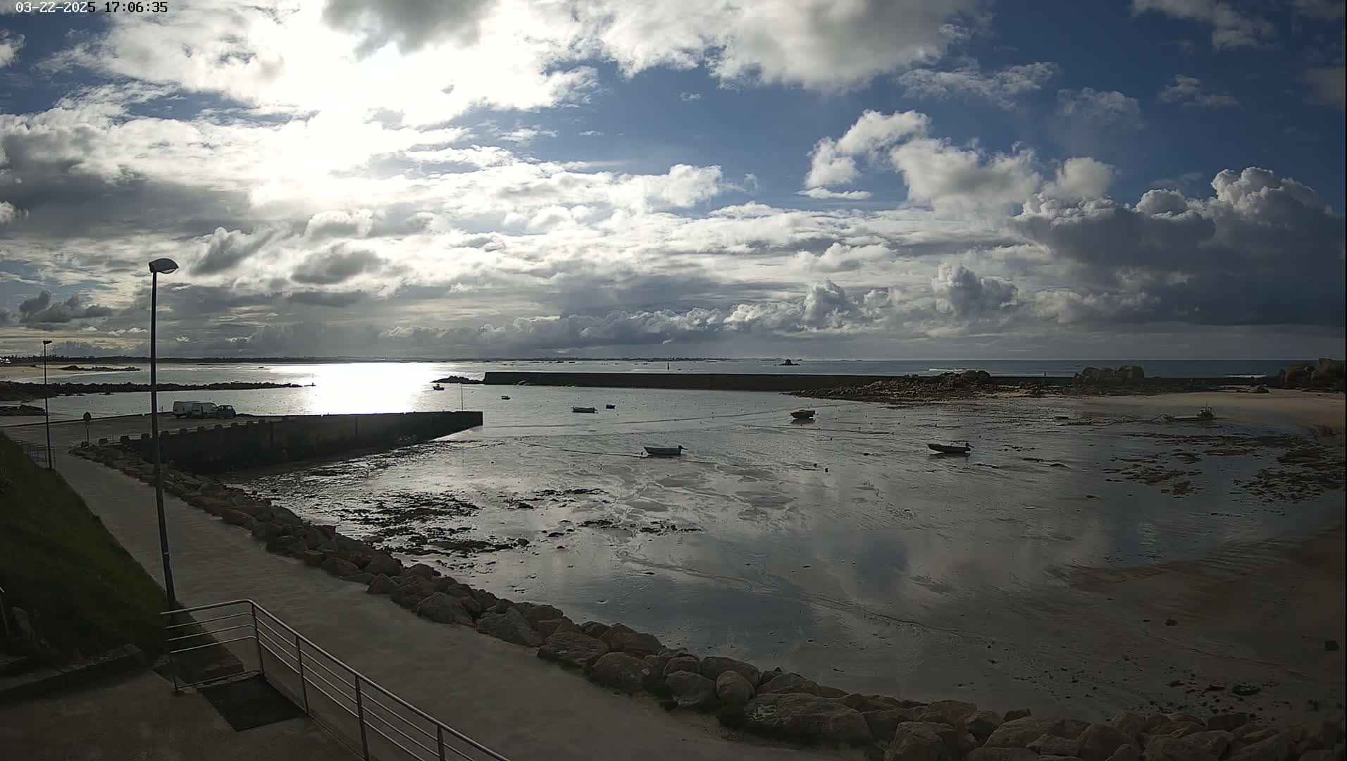 A partly cloudy sky over a calm harbor with several small boats at low tide, a paved walkway leading to the water's edge, and a van parked nearby.