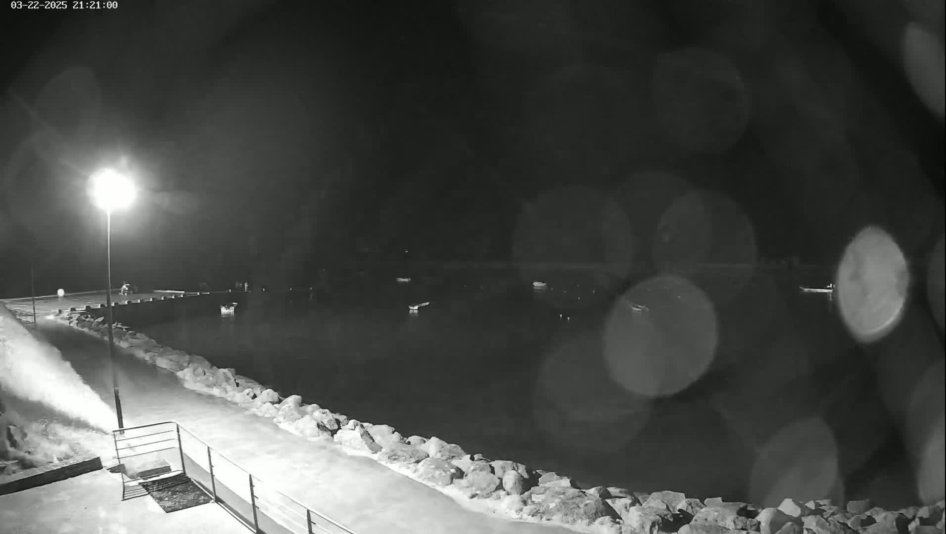 A snow blower clears snow from a nighttime waterfront area with a calm body of water containing several boats.