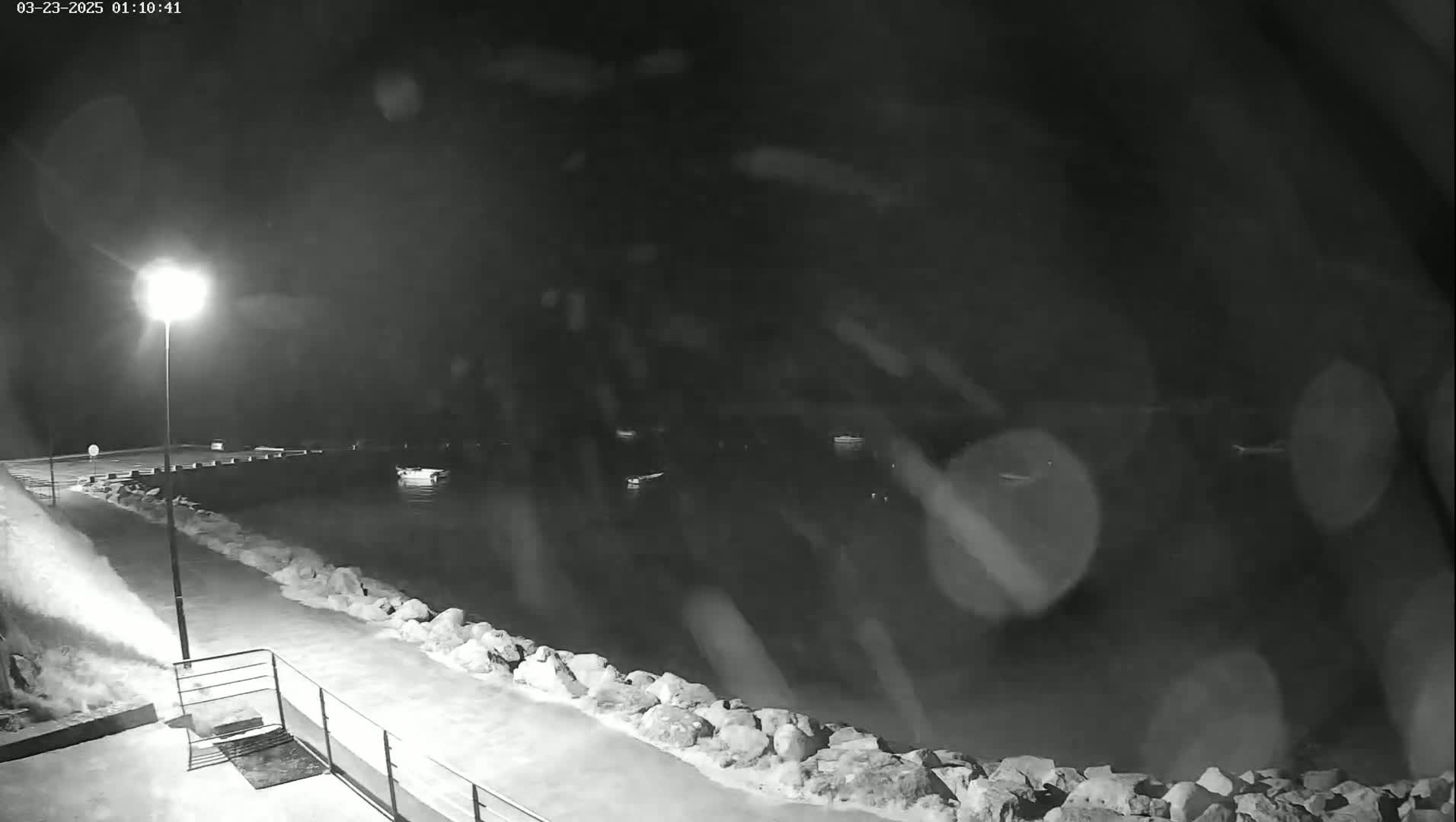 A nighttime, snowy scene shows a rocky shoreline with small waves lapping against it, illuminated by a single lamppost, with a few boats visible in the dark water beyond.