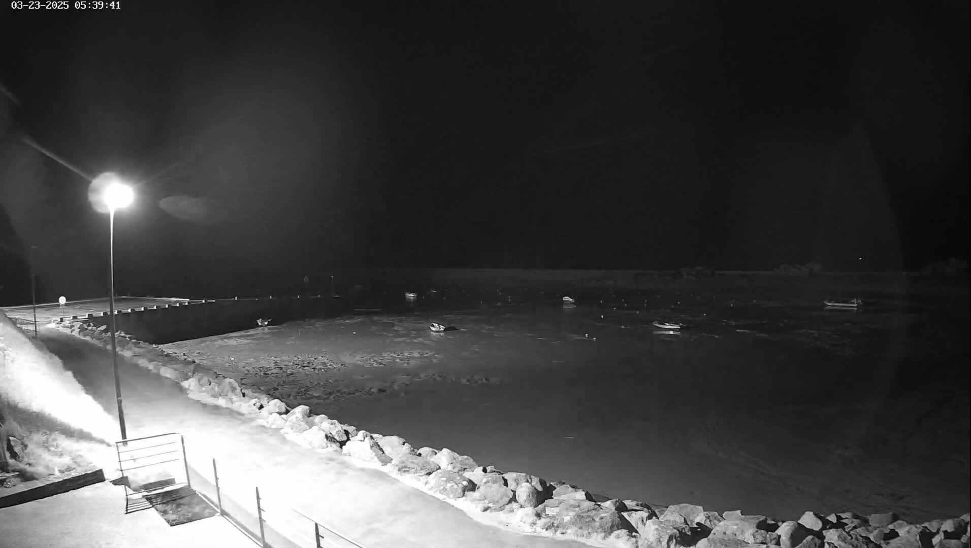 A nighttime, snow-covered scene shows a calm body of water with several small boats moored, viewed from a snow-covered rocky shore lit by a single lamppost.