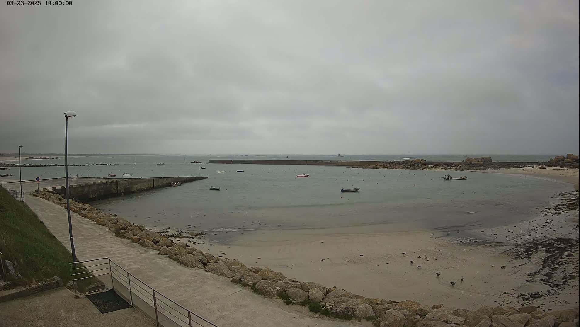 A calm, overcast day reveals a small harbor with several boats, a sandy beach, and rocky outcroppings.