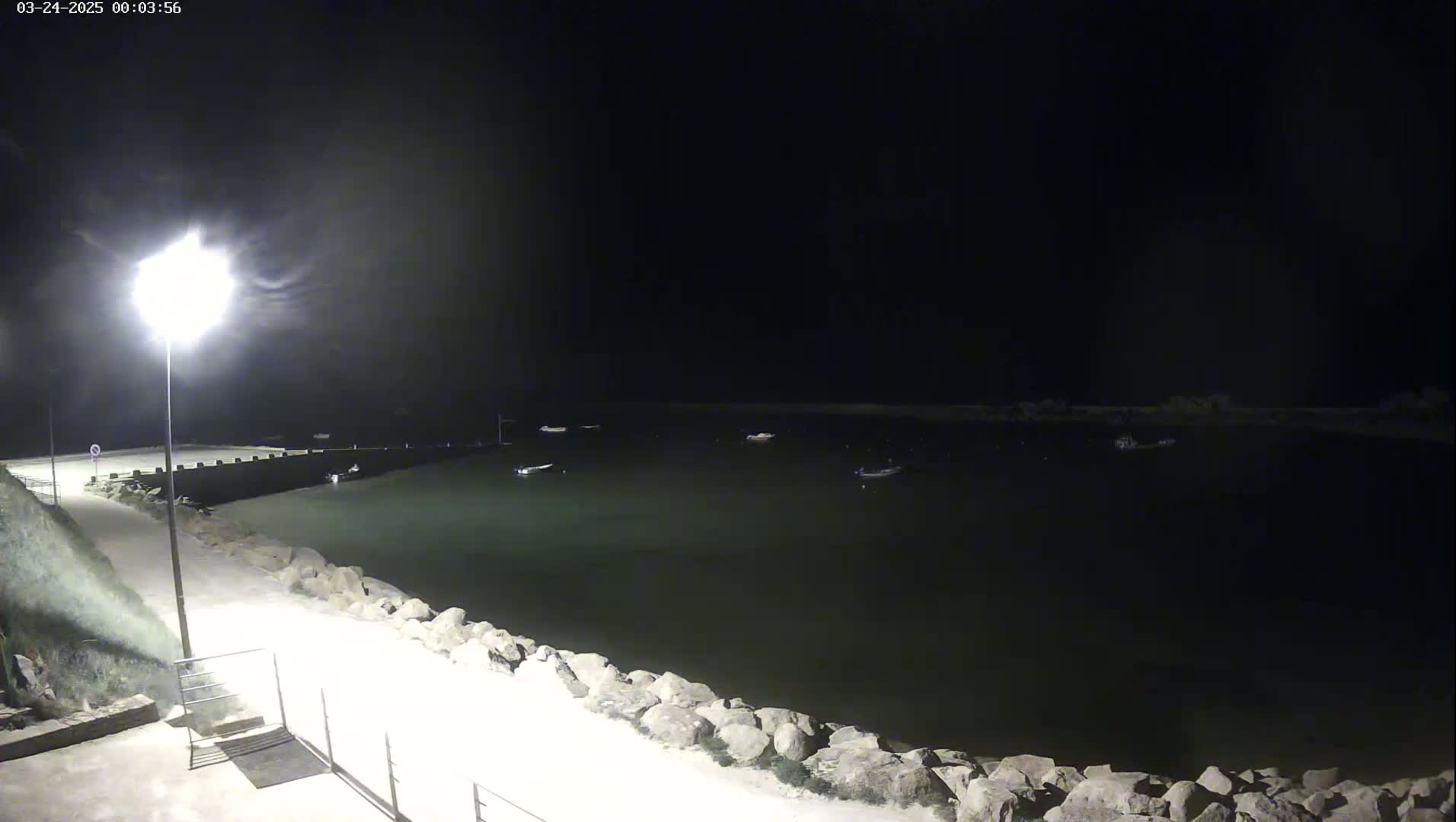 A nighttime view of a calm harbor with several boats at anchor, illuminated by a single bright lamppost along a paved walkway beside a rocky embankment.