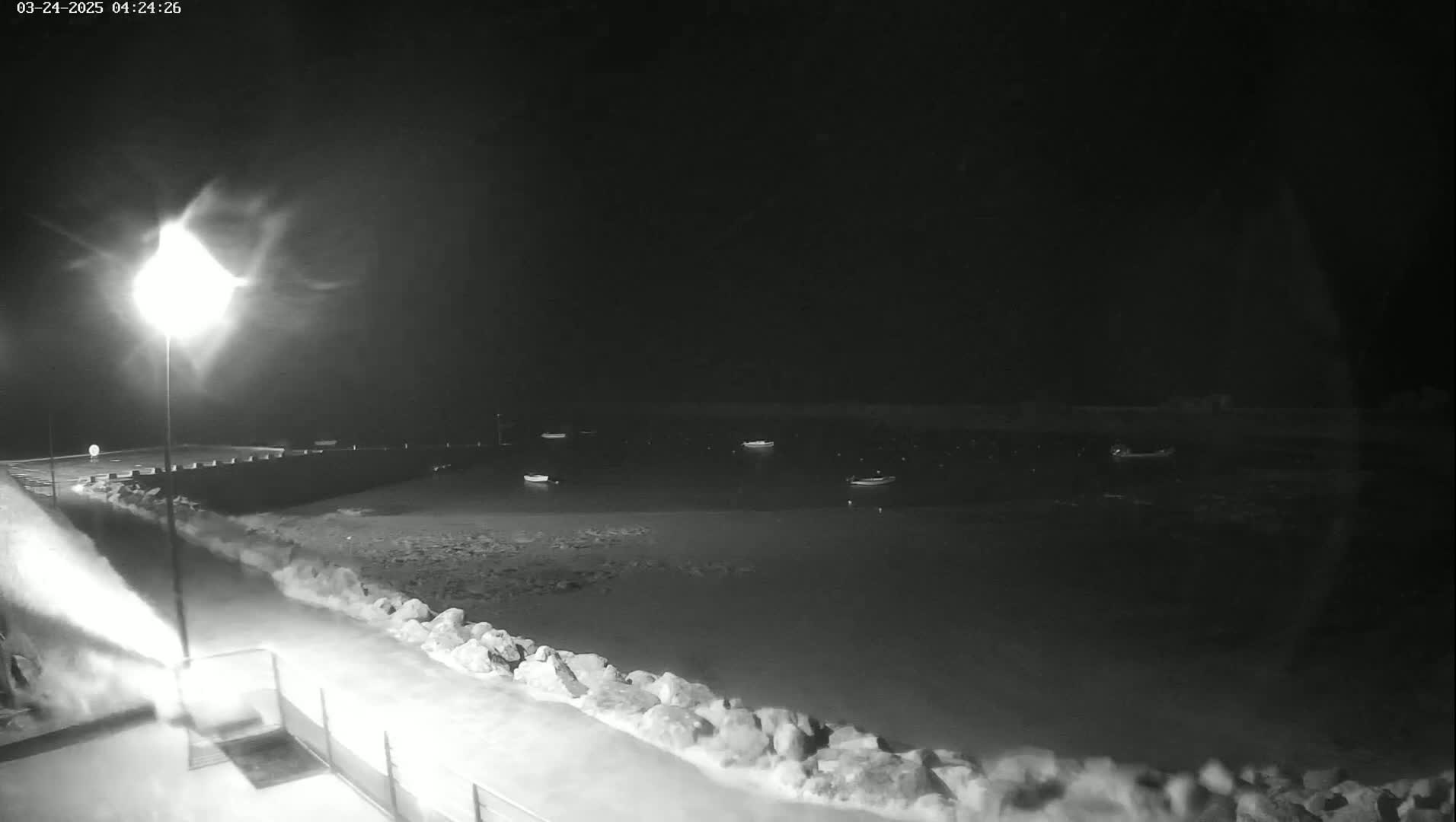 A nighttime view of several small boats moored in calm water next to a snow-covered rocky shoreline, illuminated by a bright streetlight.