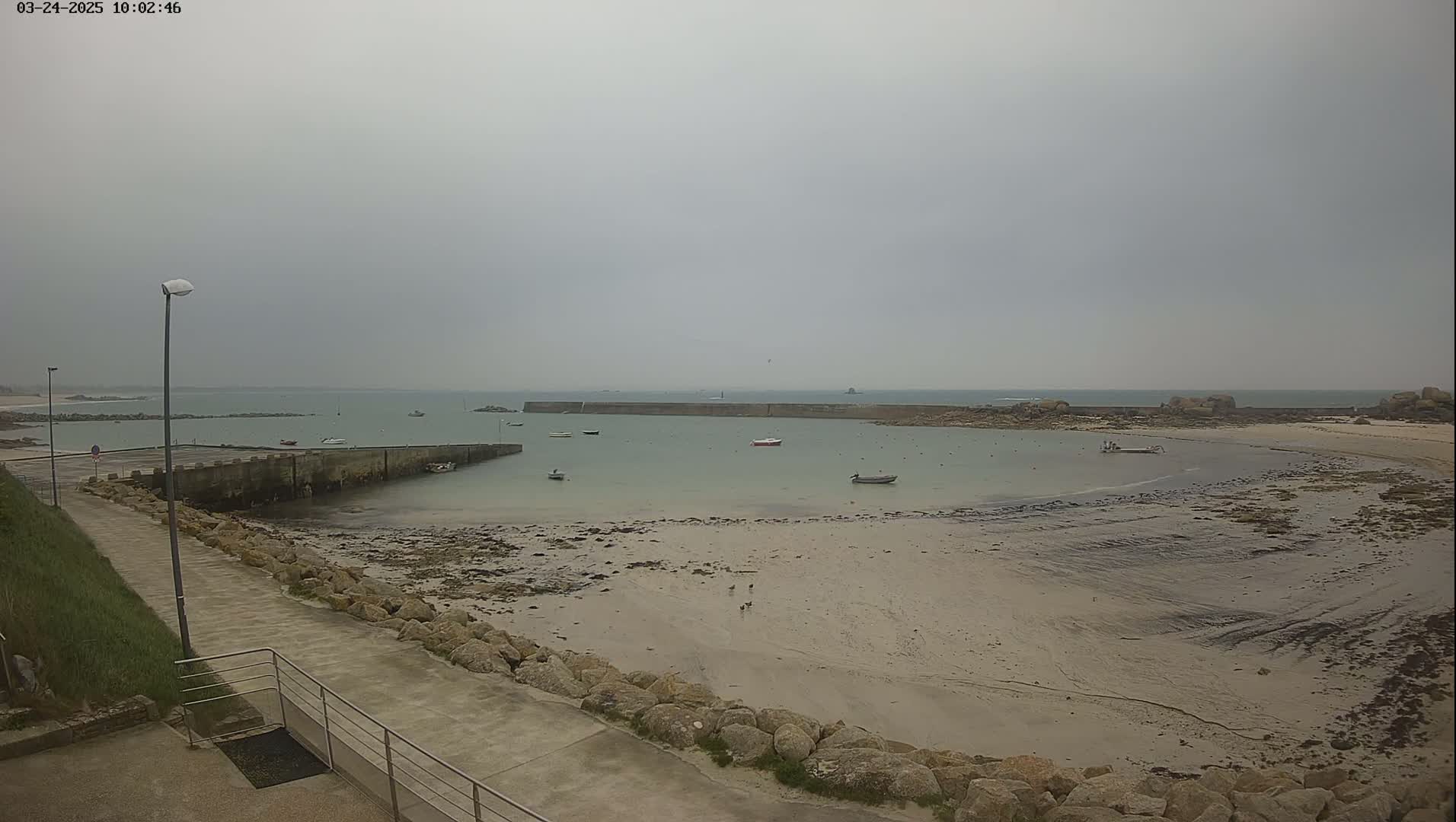 A calm, overcast day shows a sandy beach with a few small boats in the water near a stone wall and a paved walkway.