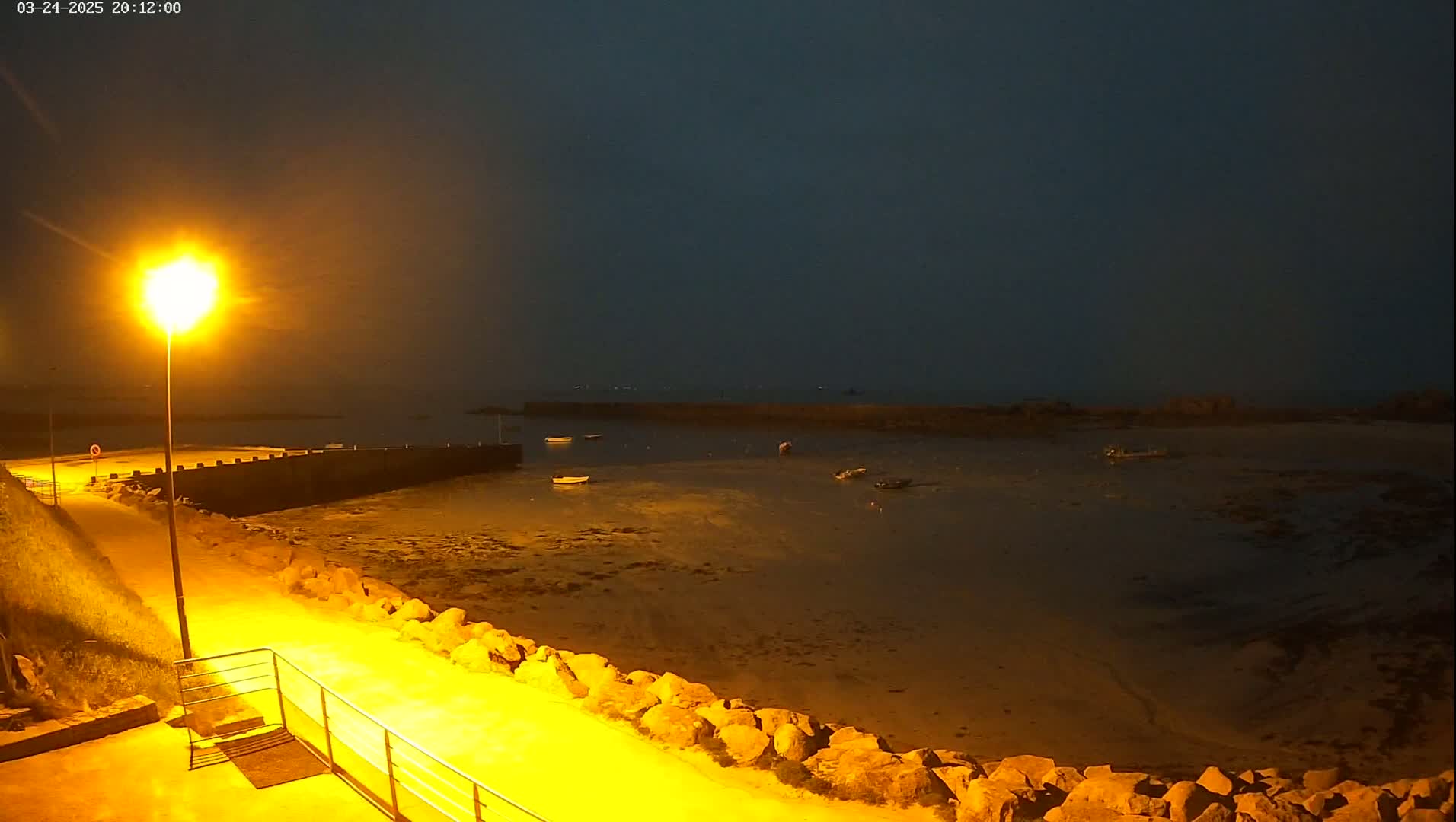 A nighttime view of a calm, shallow harbor with several small boats at low tide, illuminated by a bright lamppost on a paved walkway.