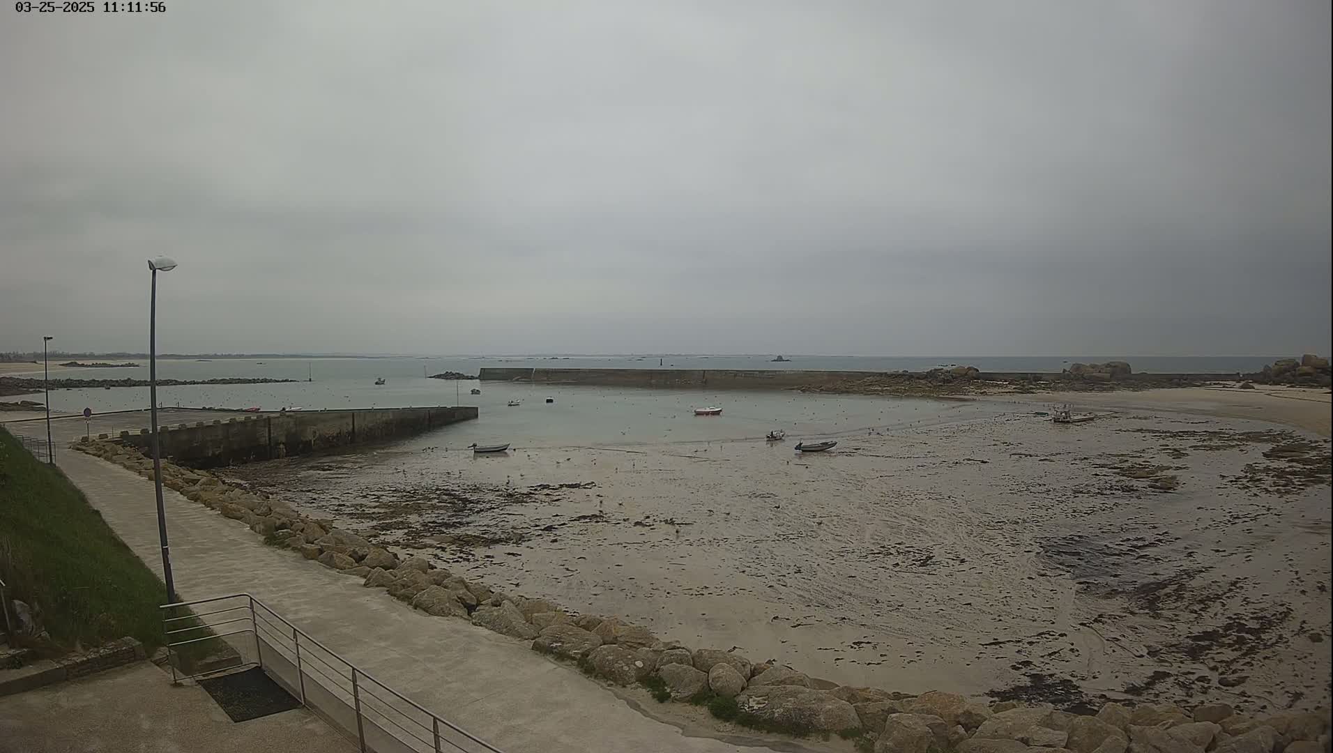 A mostly overcast day reveals a harbor with several small boats at low tide, a concrete walkway, and a rocky shoreline extending to a calm sea.