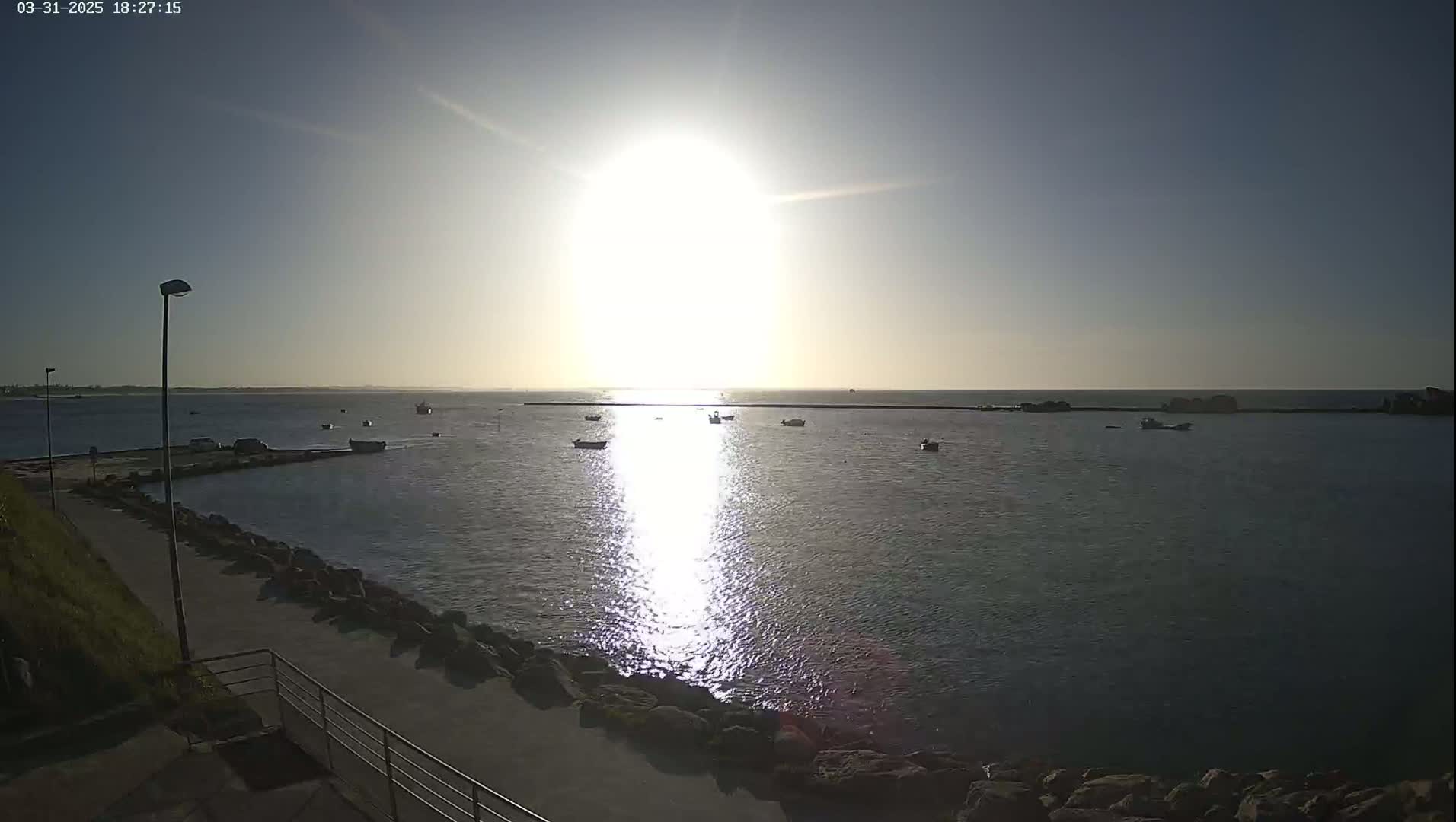 A bright sun shines over a calm body of water with several small boats, viewed from a paved walkway next to a rocky shoreline.