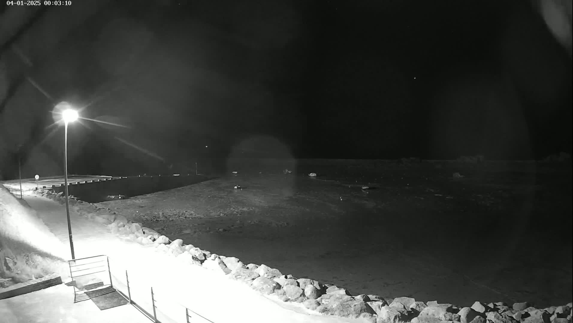 A nighttime, snow-covered waterfront scene is illuminated by a single lamppost, showing a calm body of water with several boats, and a snow-covered walkway.