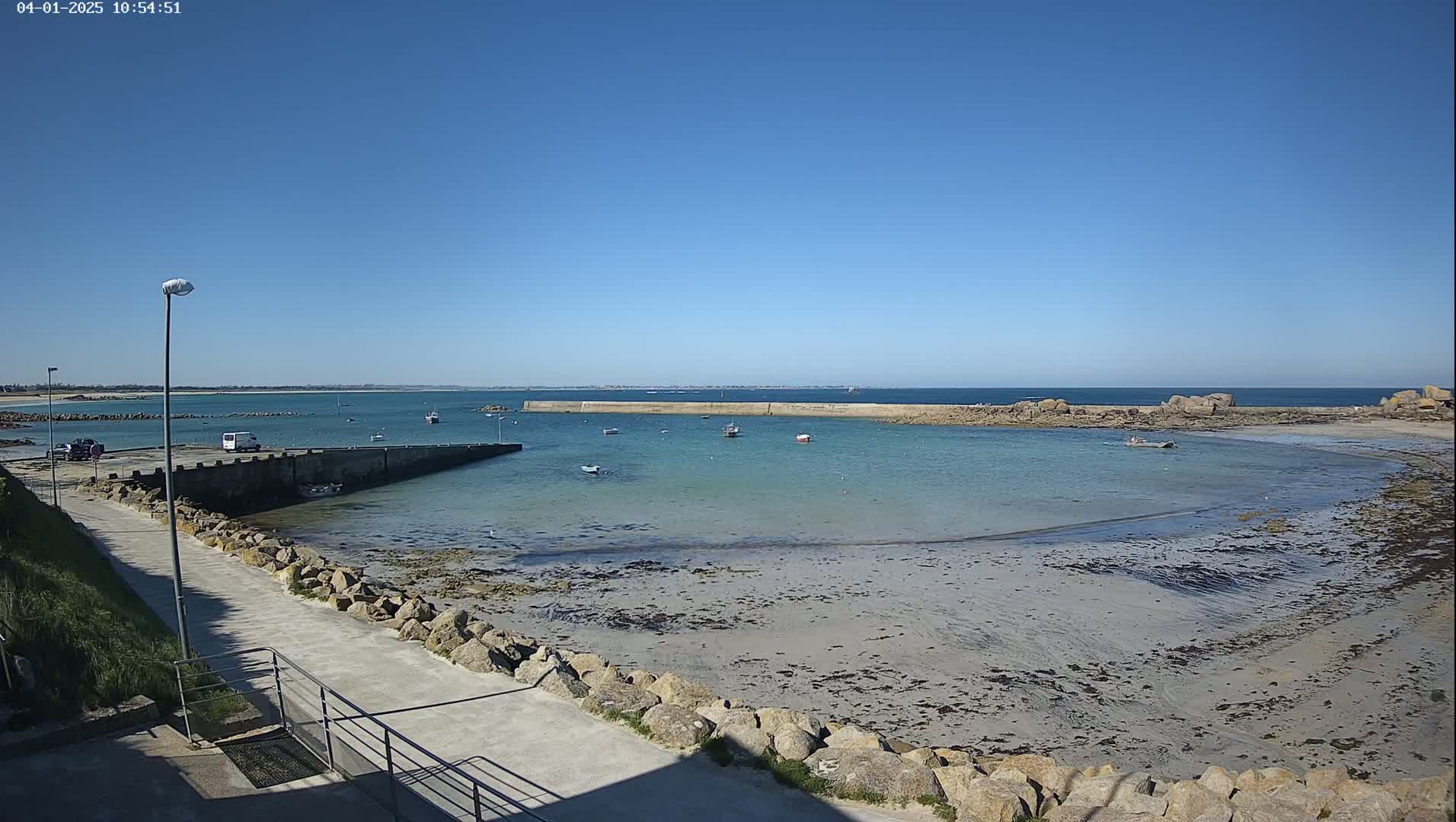 A calm, sunny day reveals a small harbor with several boats, a sandy beach, and a rocky shoreline.