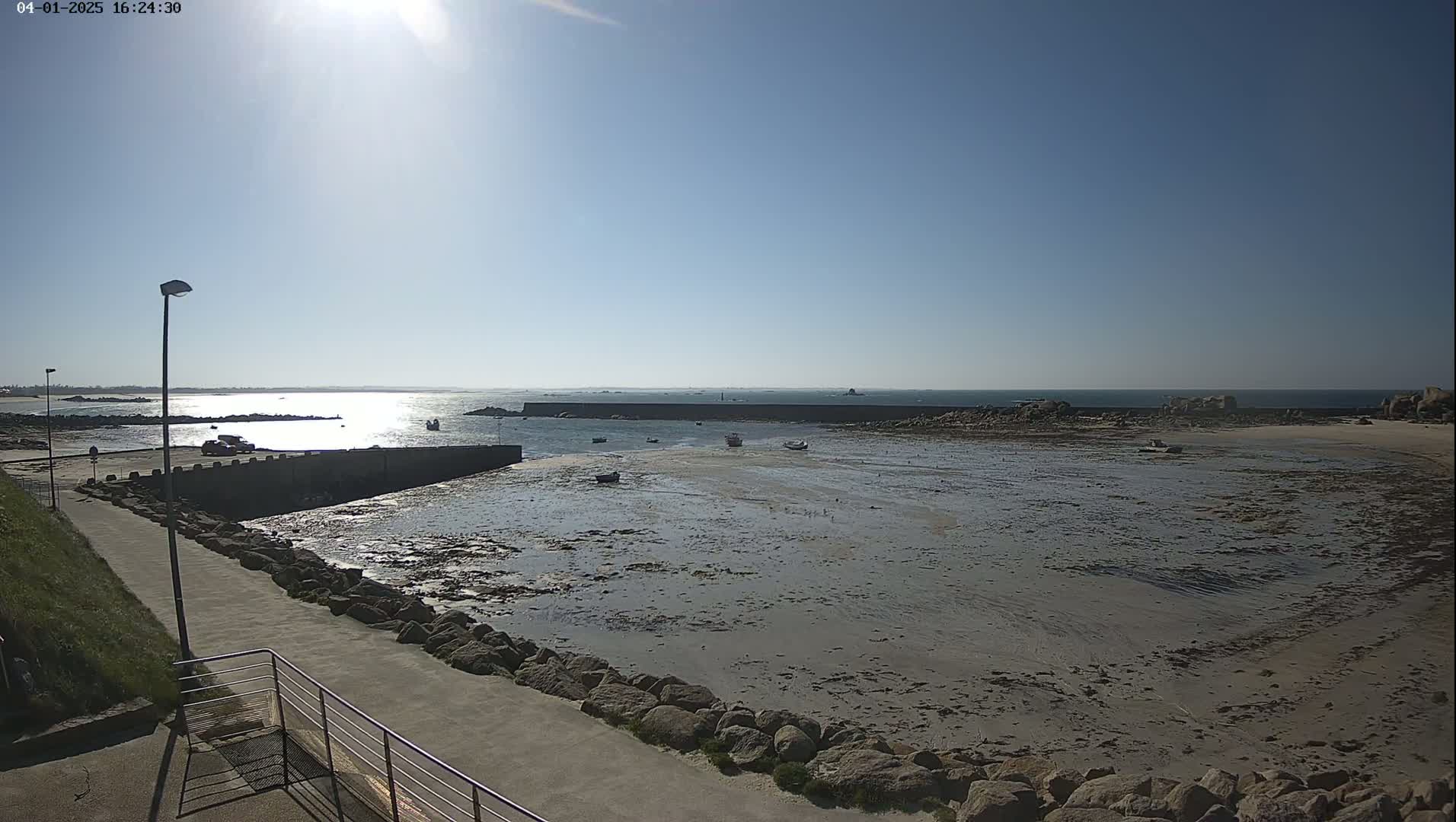 A sunny day reveals a mostly exposed harbor with several small boats, a low sea wall, and a paved walkway leading to it.