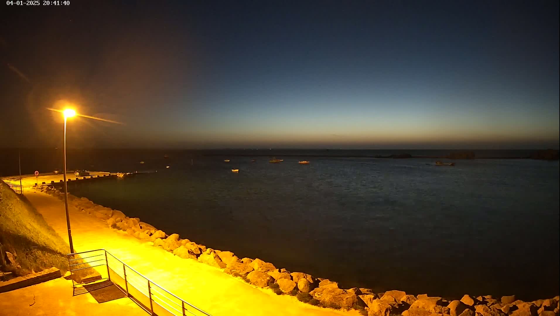 A paved walkway lit by a lamppost overlooks a calm body of water at dusk, where several small boats are moored.